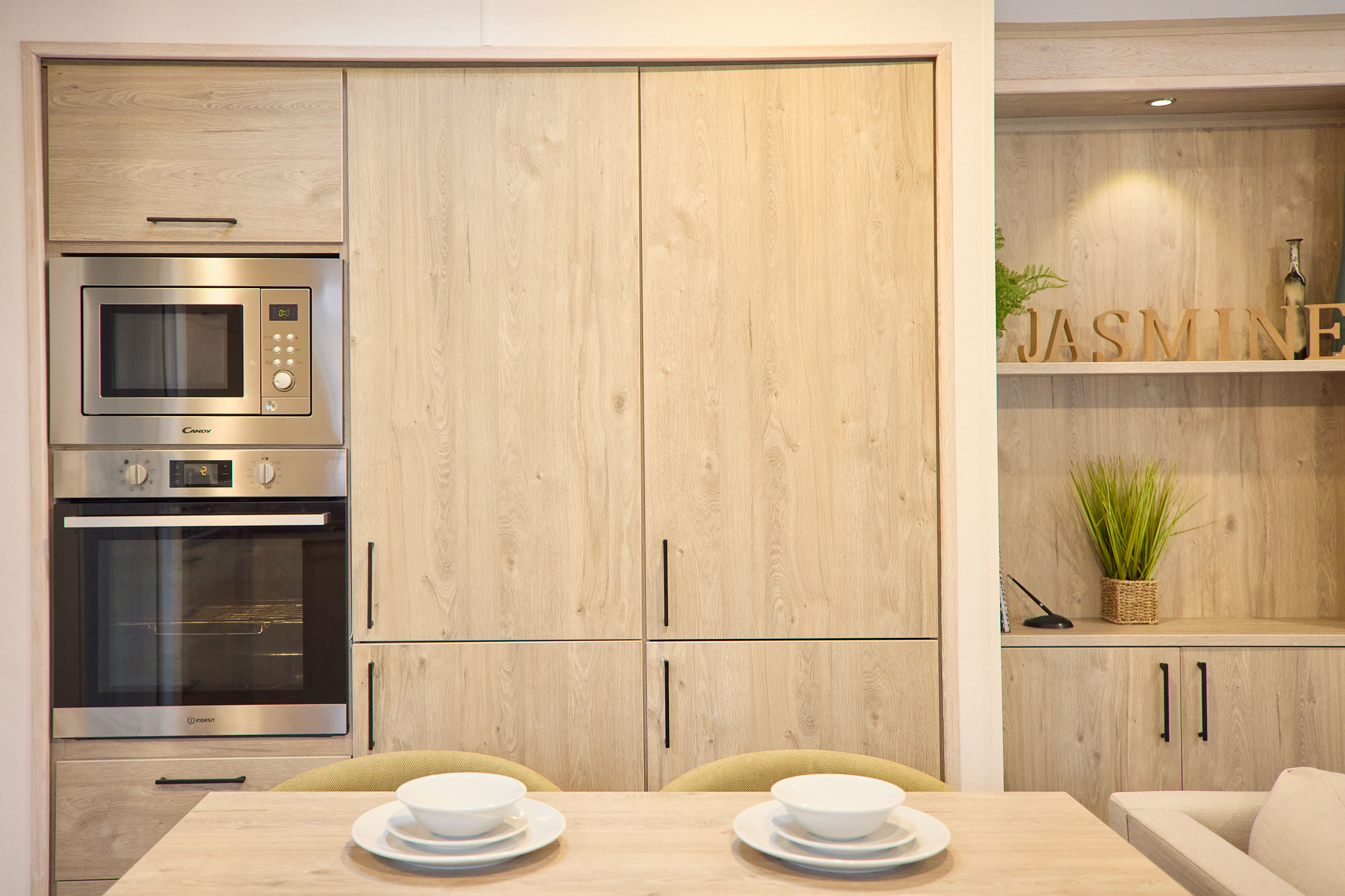 Close-up view of the modern kitchen's light wood cabinetry, featuring integrated stainless steel microwave and oven units. A dining table with place settings is visible in the foreground.