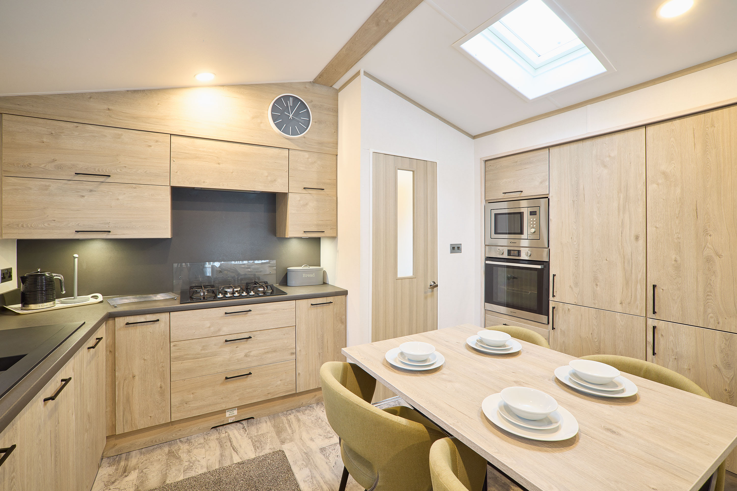 Modern kitchen and dining area featuring light wood cabinetry, dark grey countertops, integrated appliances, and a dining table set for four under a bright skylight.