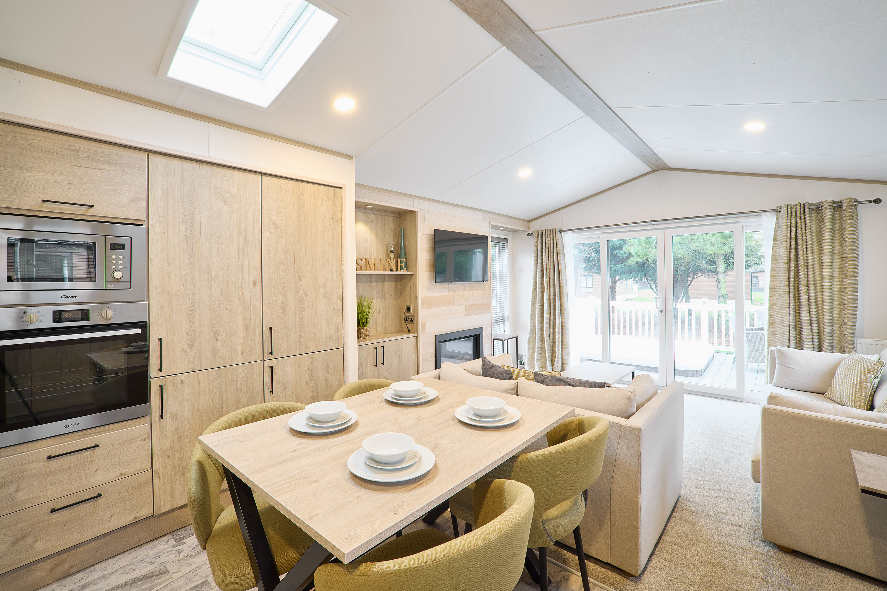 Spacious open-plan area featuring a modern kitchen with integrated appliances, a dining table set for four, and a comfortable sofa. Sliding glass doors provide access to an outdoor deck with a hot tub.