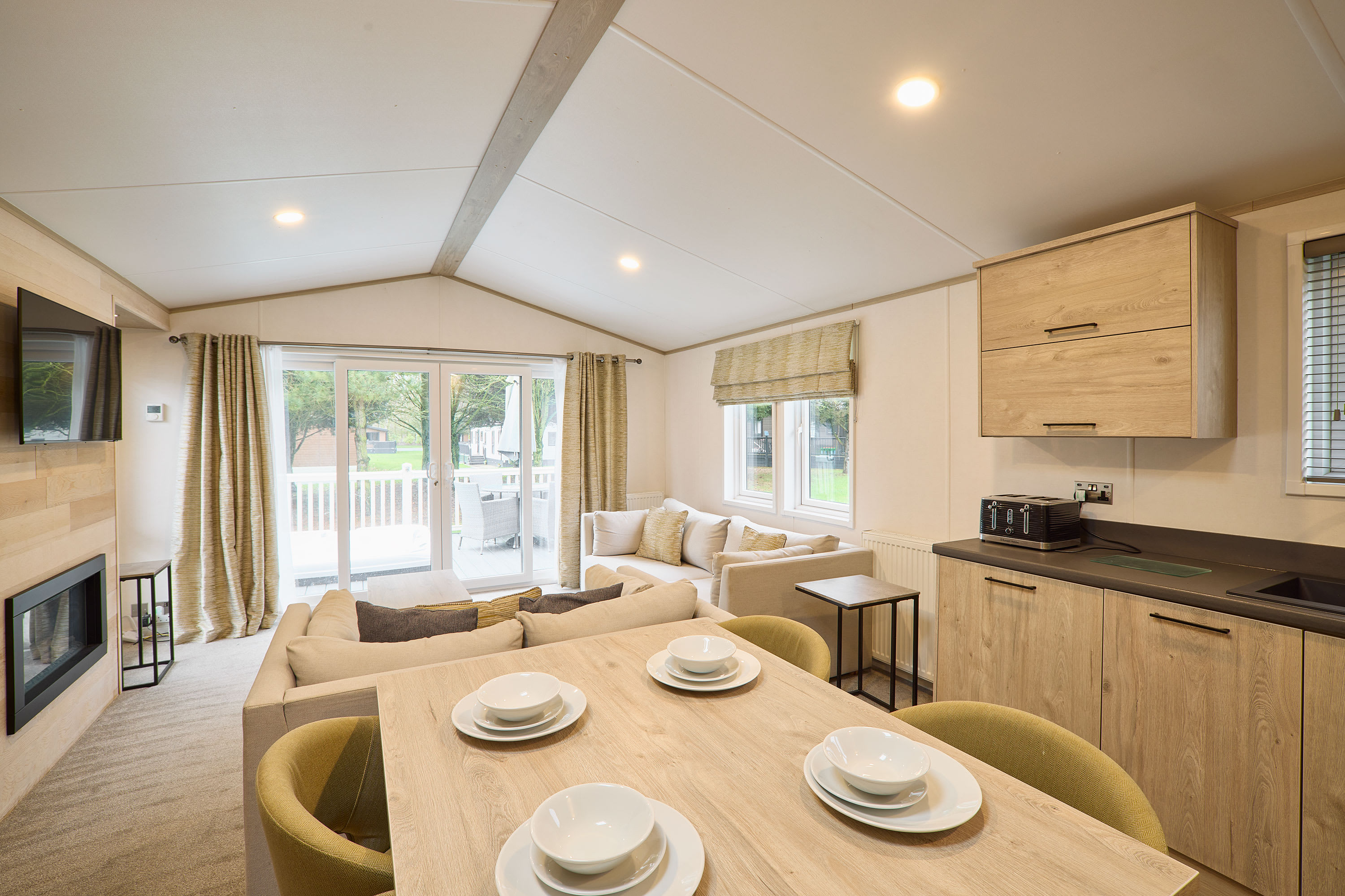 Bright open-concept living and dining area with a light-colored sofa, a wooden dining table set for four, and a partial view of the kitchen. Large sliding glass doors lead to an outdoor deck.