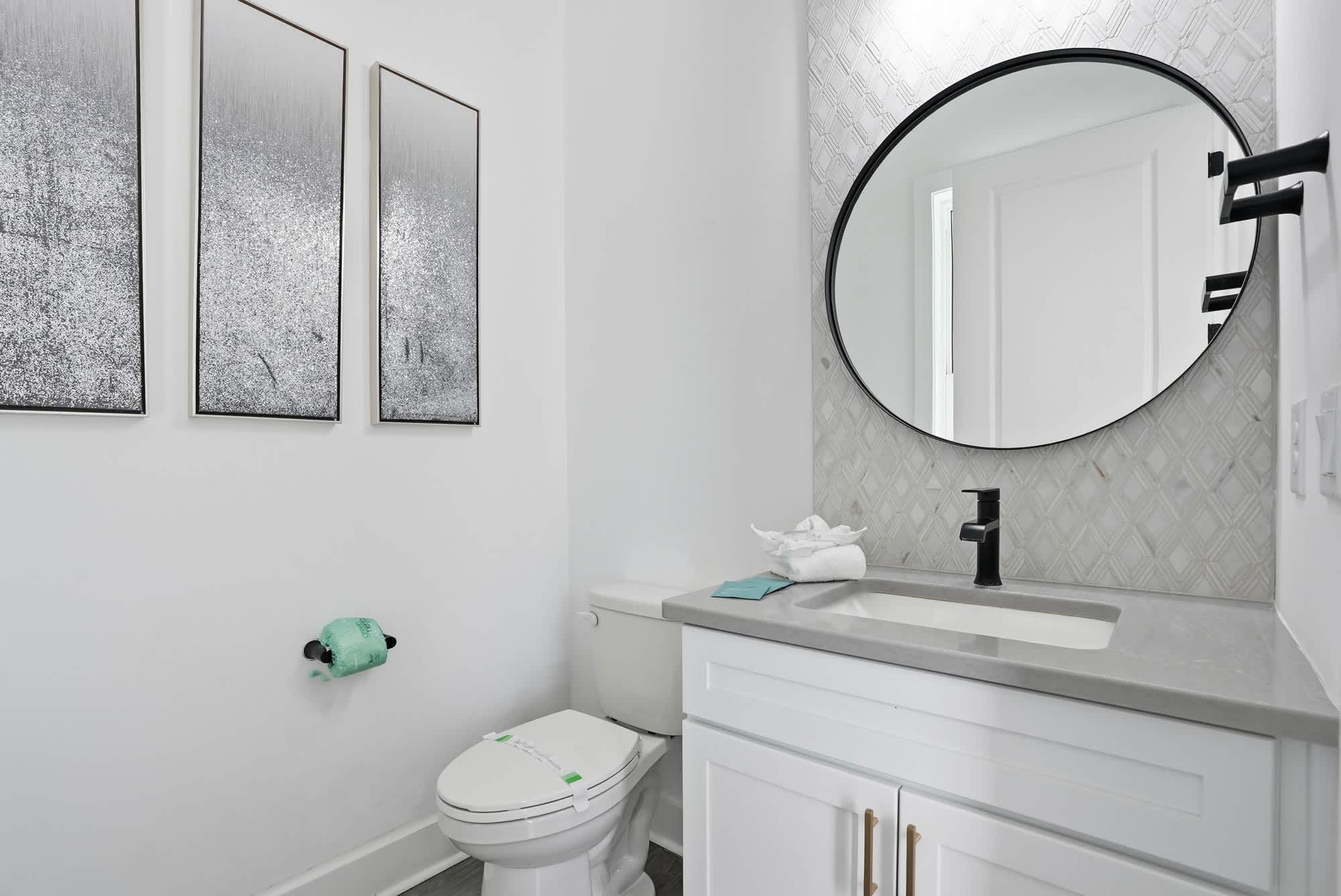 120 Dune Drive – Modern bathroom with circular mirror, gray countertop, white cabinetry, and three framed artworks on the wall.
