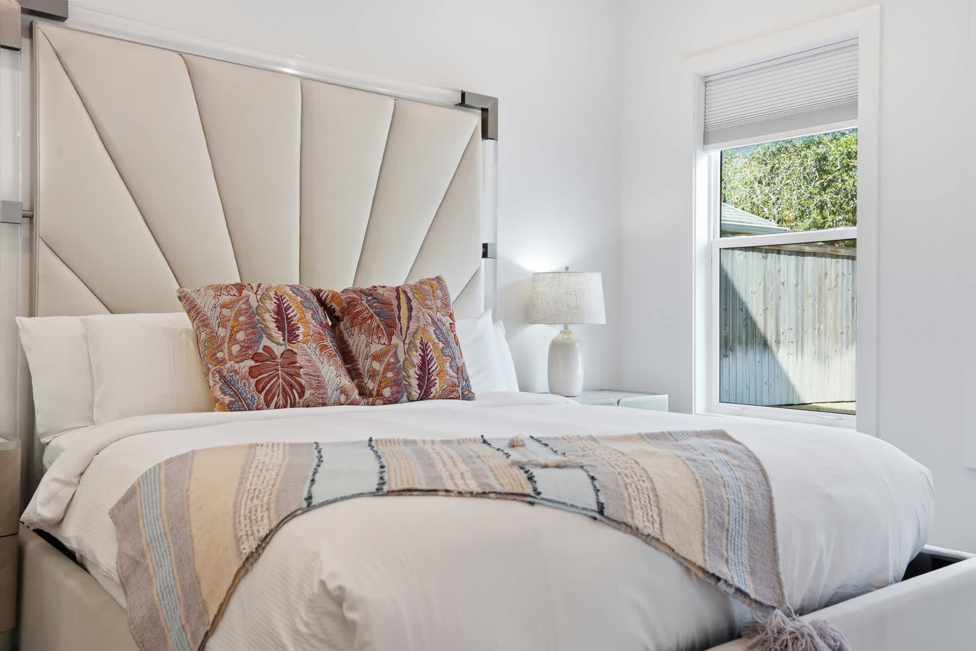 120 Dune Drive – Bright bedroom with a plush headboard, decorative pillows, bedside lamp, and window view of a wooden fence.