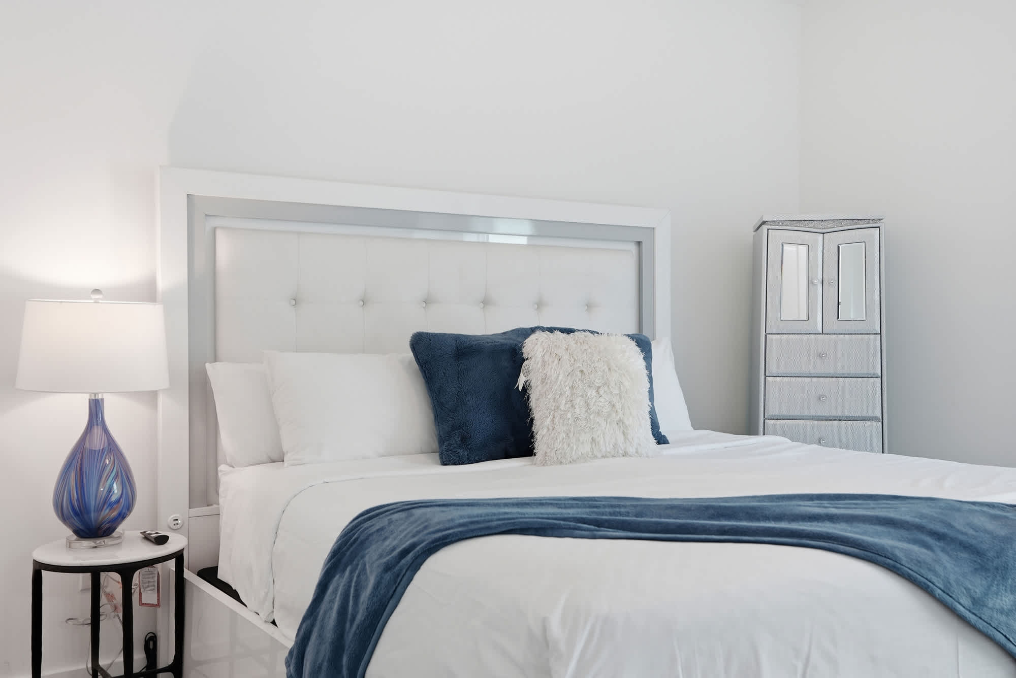 120 Dune Drive – Modern bedroom with white bed, decorative pillows, blue throw blanket, lamp, and mirrored dresser.