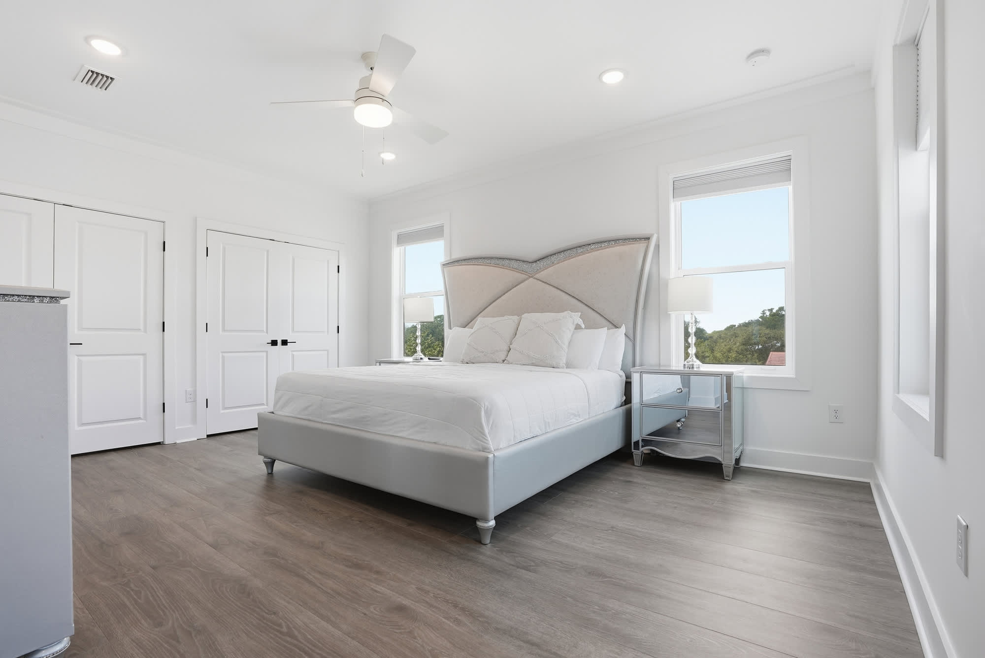 120 Dune Drive – Bright bedroom with a queen bed, two windows, and a nightstand, featuring white walls and wood flooring.