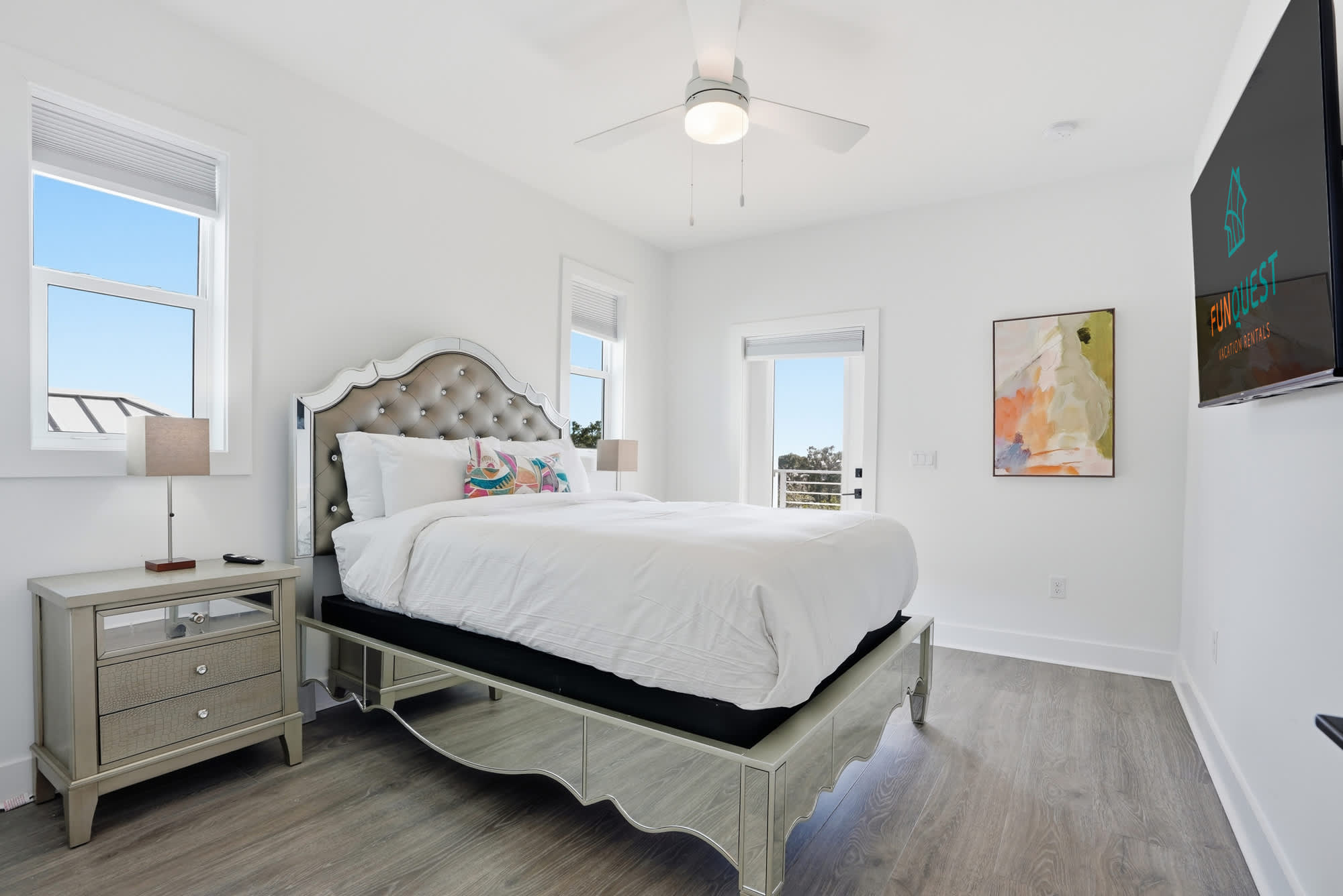 120 Dune Drive – Bright bedroom with a tufted silver bed, nightstand, ceiling fan, and two windows showcasing a view.