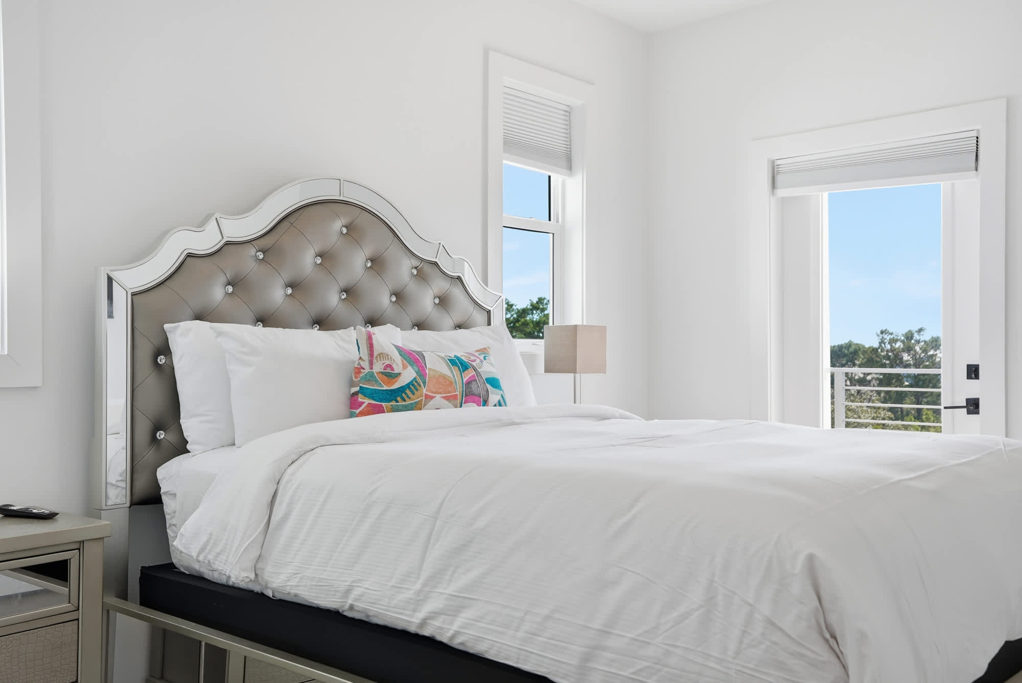 120 Dune Drive – Bedroom with a tufted headboard, white bedding, lamp, and windows showing a view of greenery.