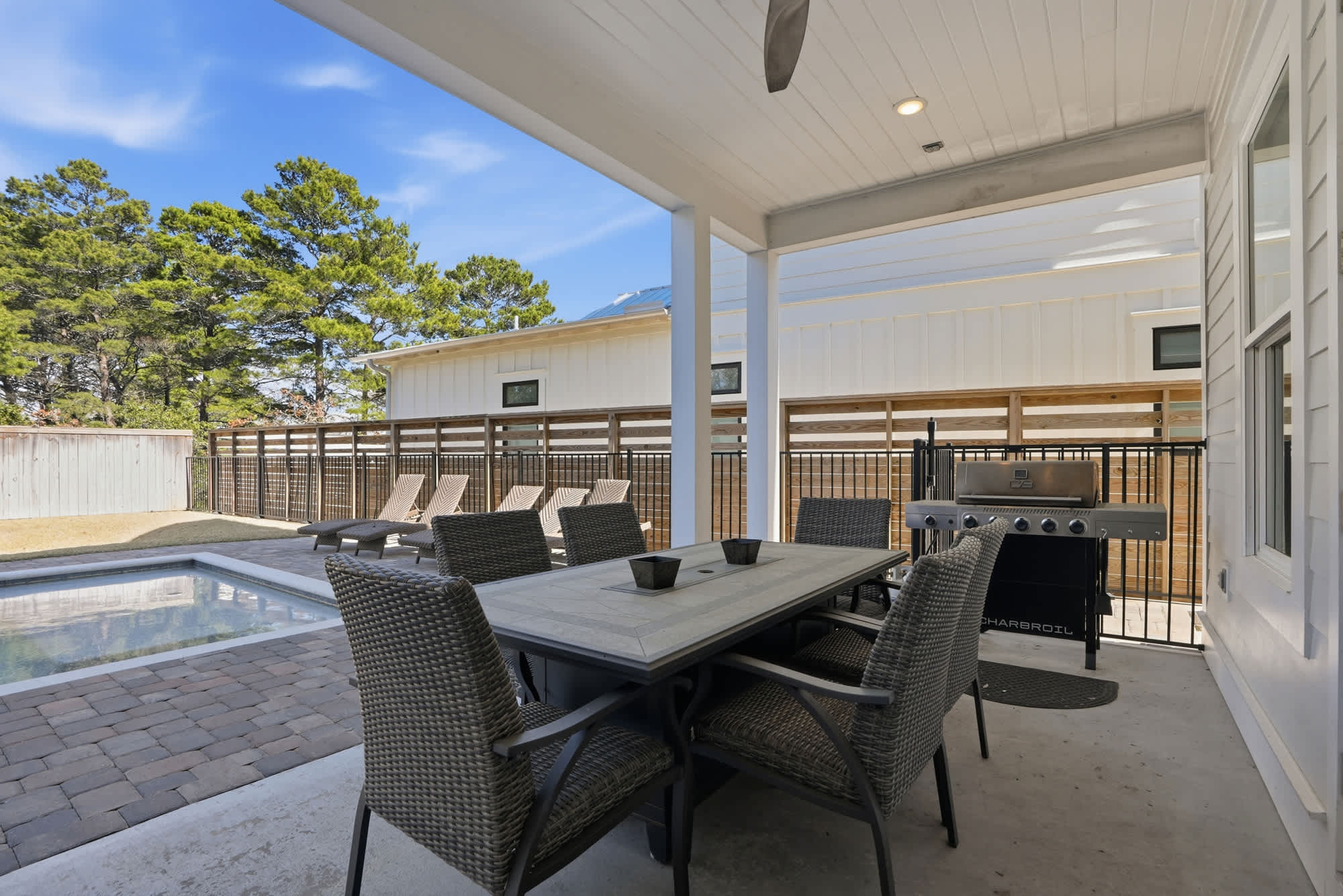 120 Dune Drive – Covered patio with dining table, chairs, grill, and pool surrounded by lounge chairs and trees.