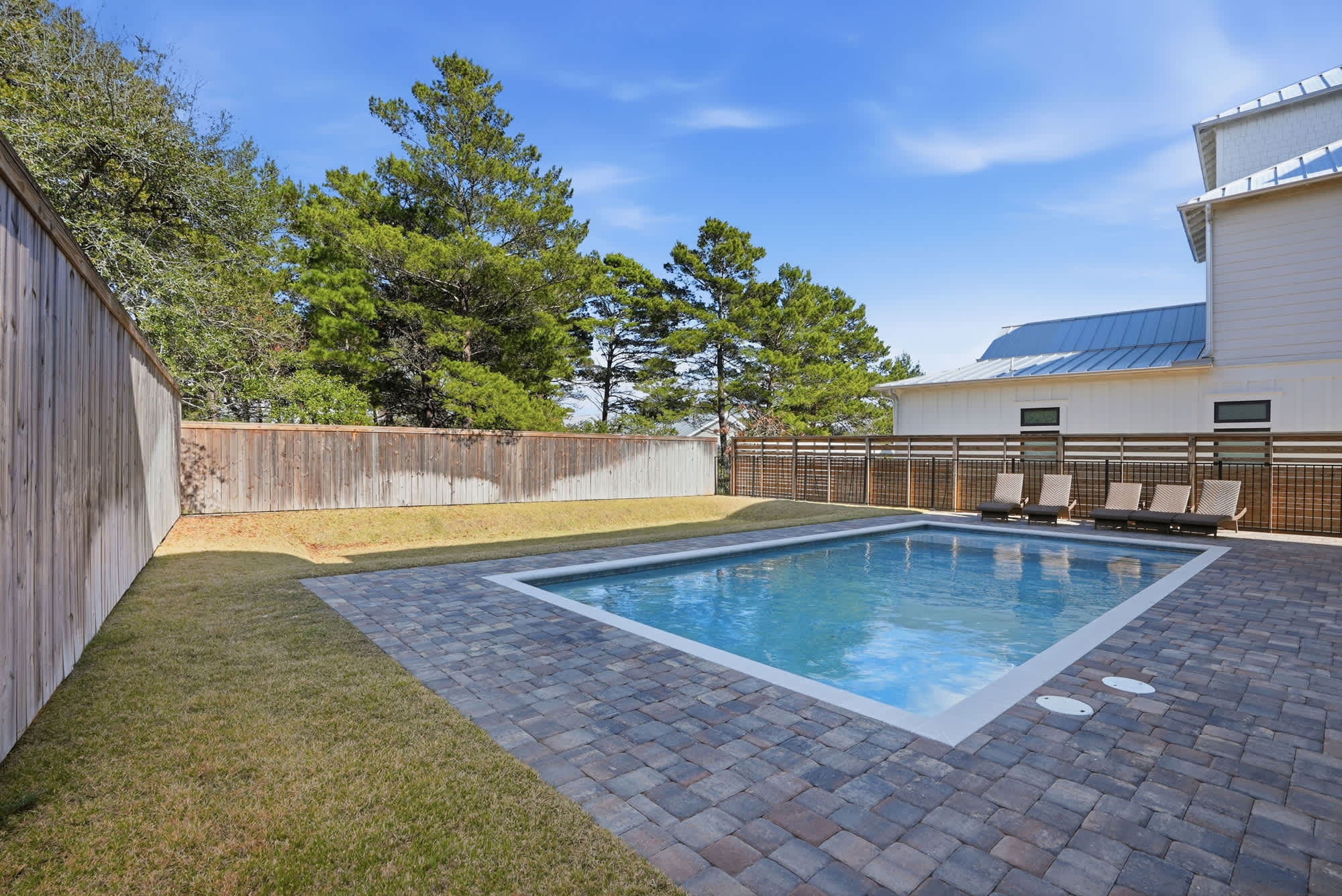 120 Dune Drive – Backyard pool area with paved patio, lounge chairs, and wooden fence, surrounded by trees and blue sky.
