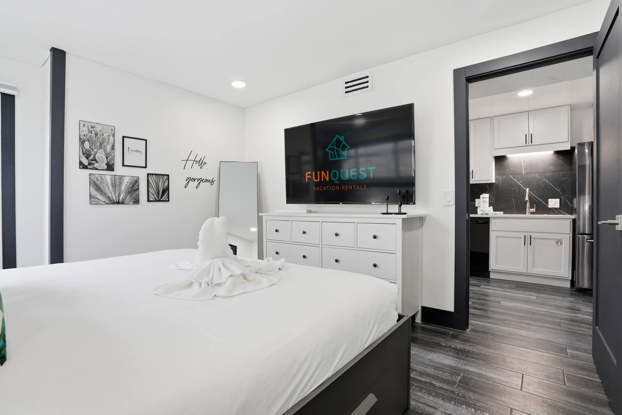 The Summit 132 – Modern bedroom with a large TV, dresser, and doorway to a kitchen area.
