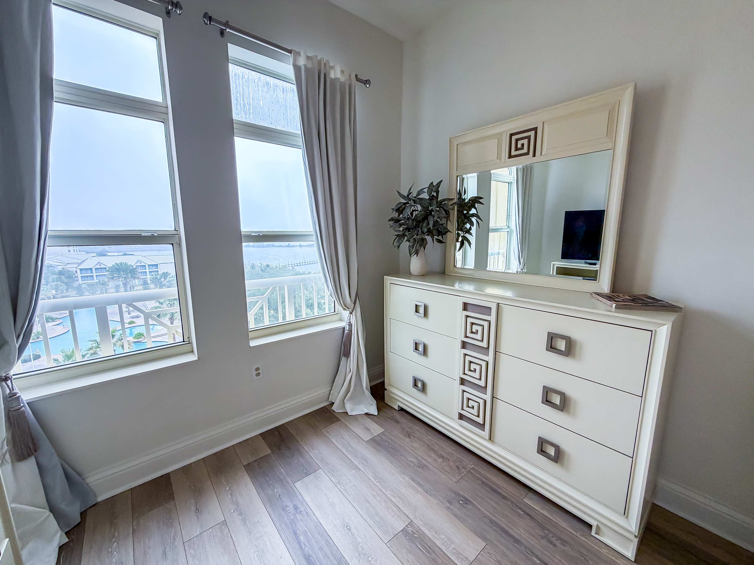 Marriott 3707 – Bedroom with large windows, dresser, mirror, and a view of a pool area and greenery outside.