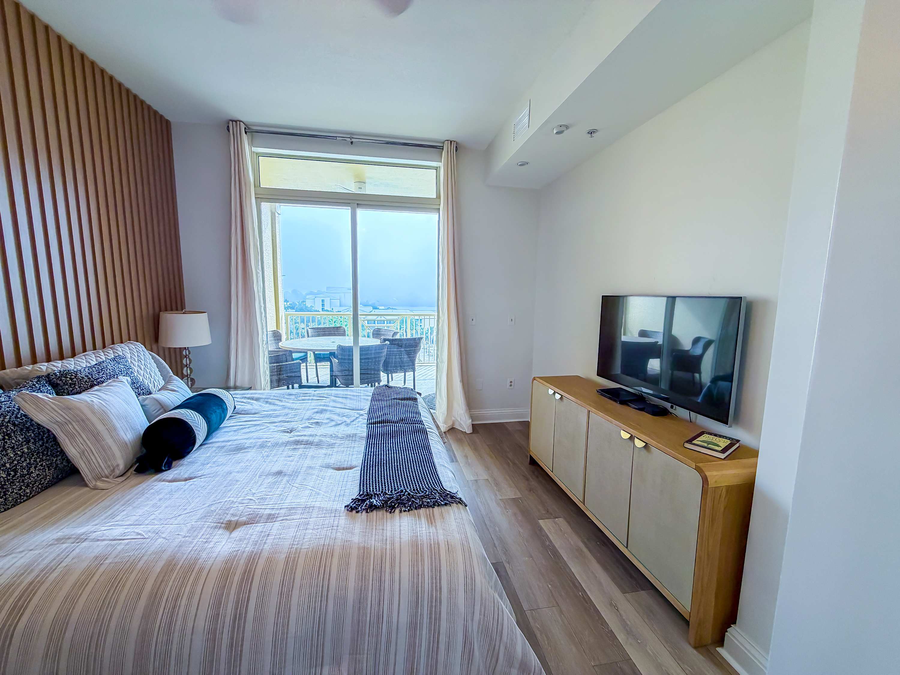 Marriott 3707 – Bedroom with a bed, large window, balcony with chairs, and a TV on a wooden console.