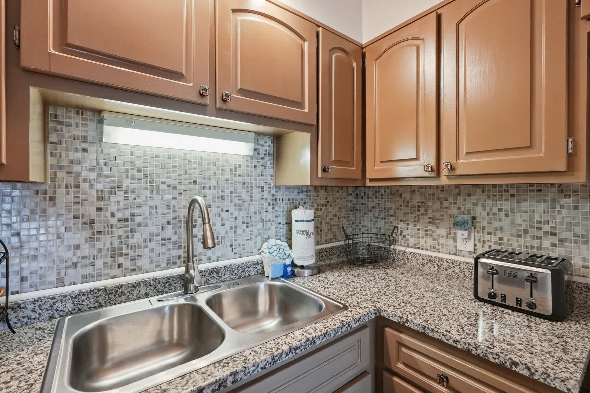 The Summit 419 – Kitchen area with stainless steel sink, granite countertops, tiled backsplash, brown cabinets, and modern toaster.