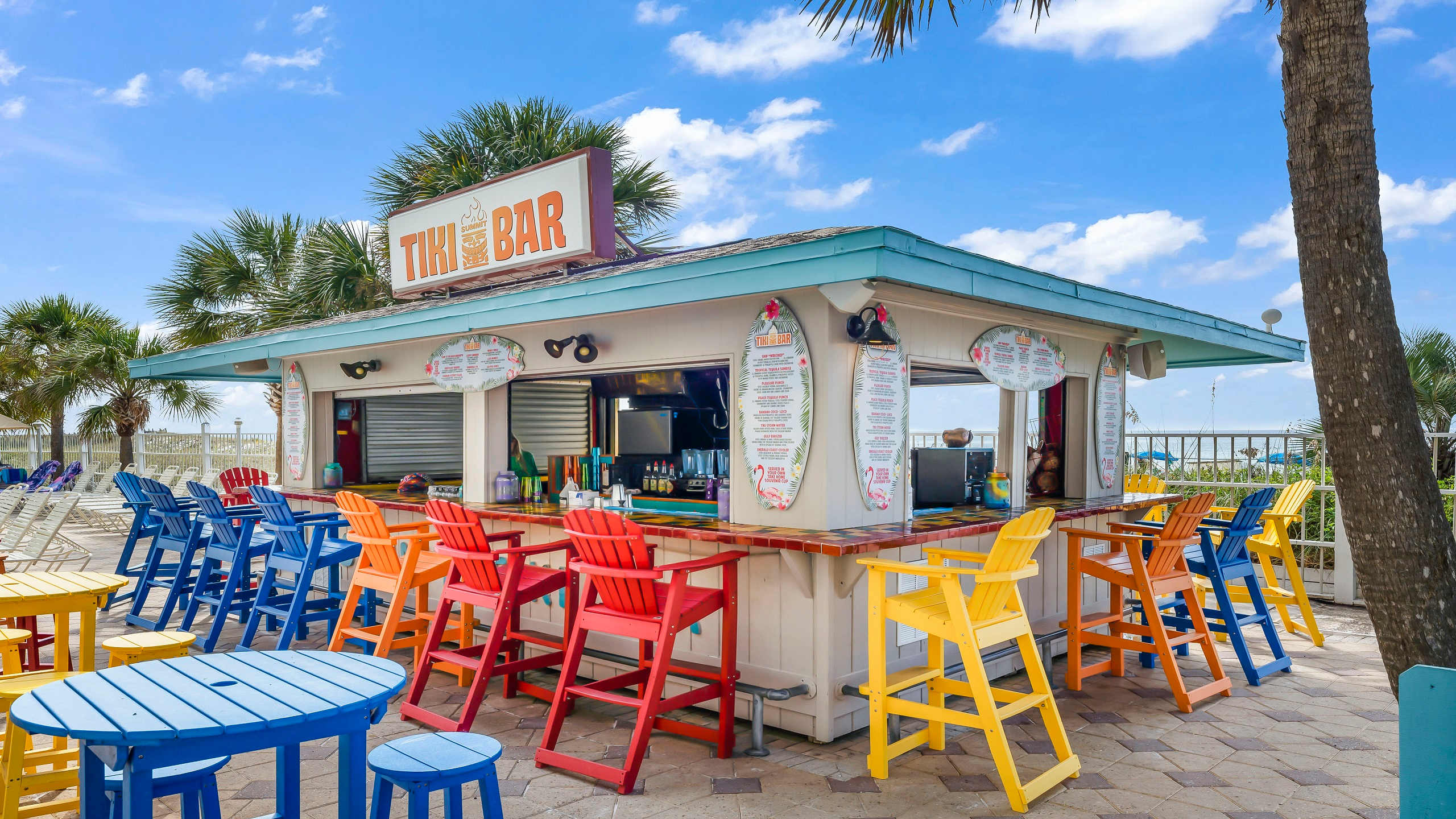 The Summit 419 – Outdoor bar with colorful high chairs, palm trees, and ocean view under a blue sky.