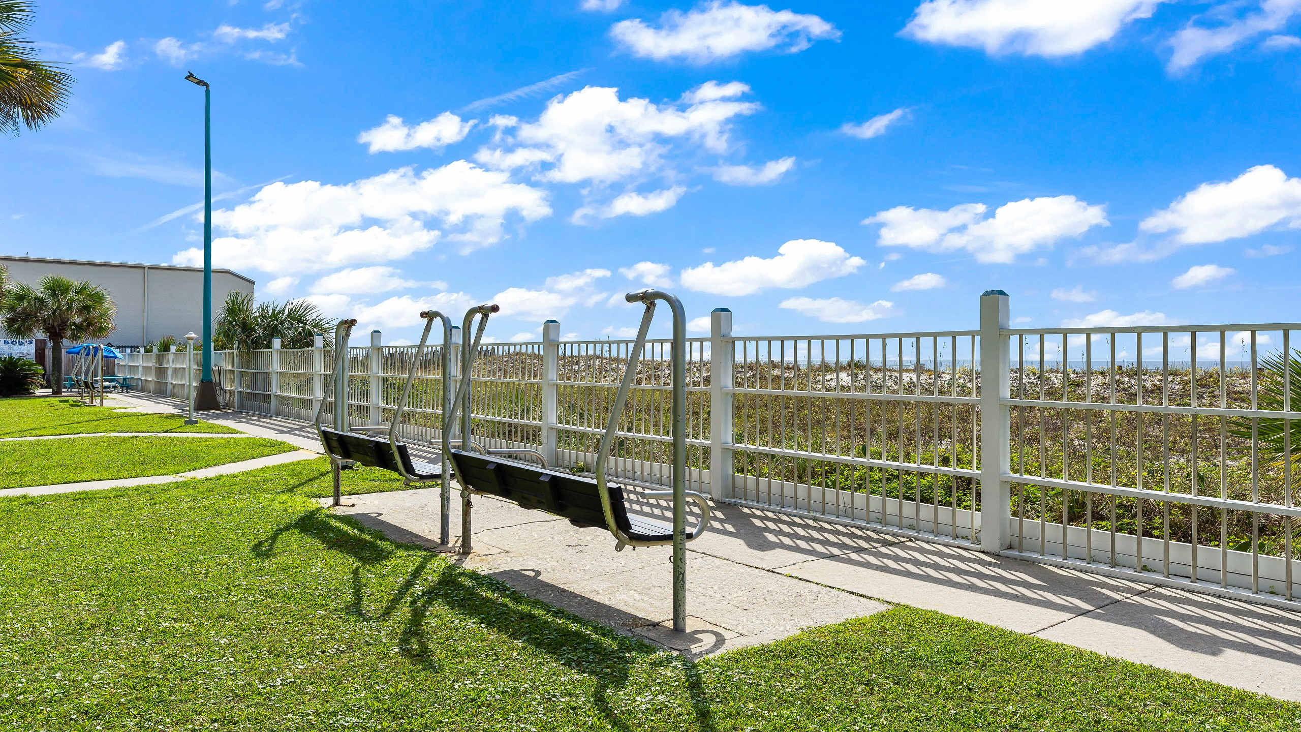 The Summit 419 – Outdoor area with swings, grassy lawn, and a view of a fence and nearby beach vegetation under a sunny sky.