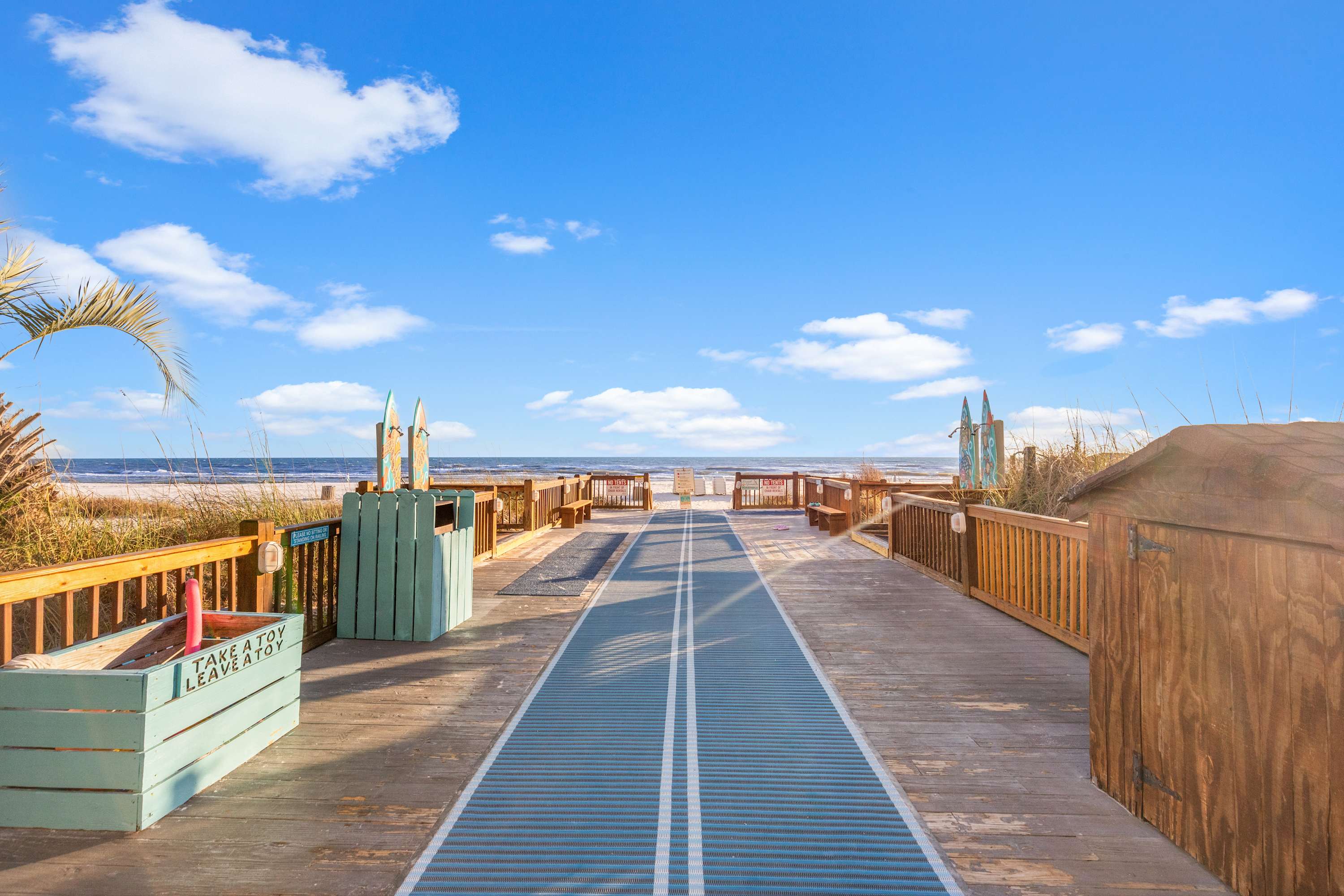 The Summit 419 – Wooden boardwalk leading to the beach with blue sky and decorative surfboards on either side.
