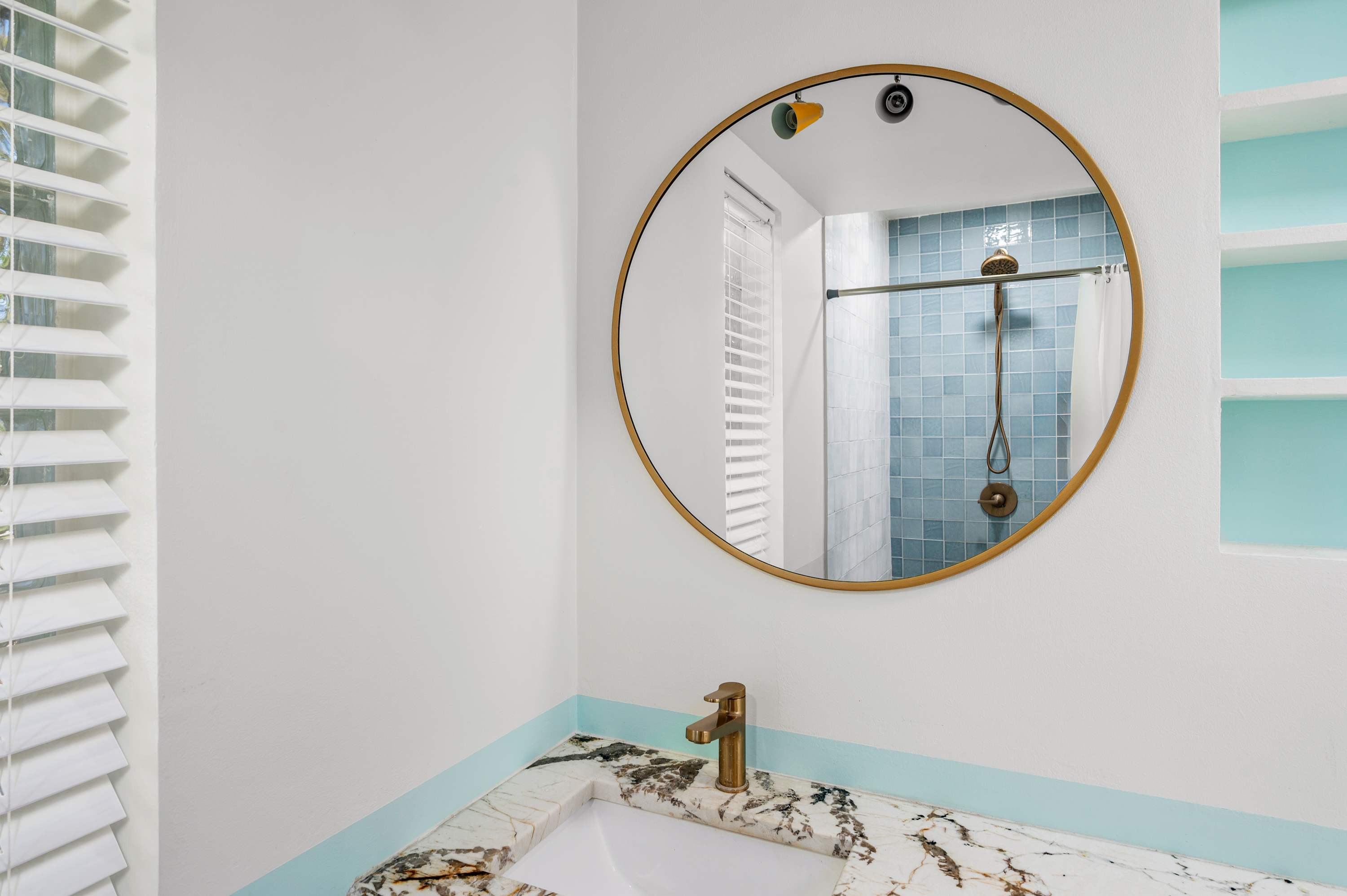 Modern bathroom sink with marble countertop and round mirror reflecting a blue-tiled shower.
