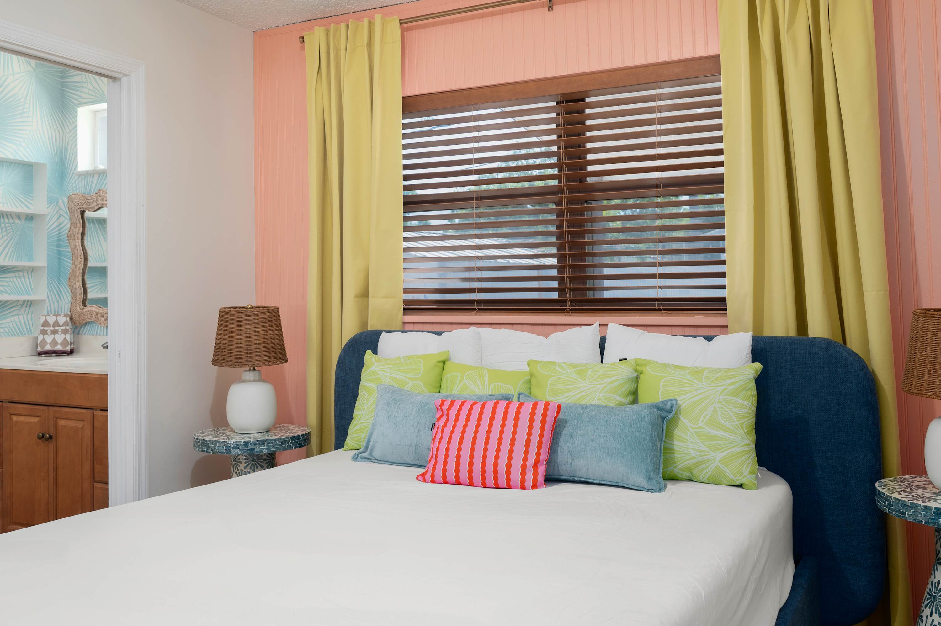 A cozy bedroom featuring a blue upholstered bed with colorful pillows and natural light from wooden blinds.