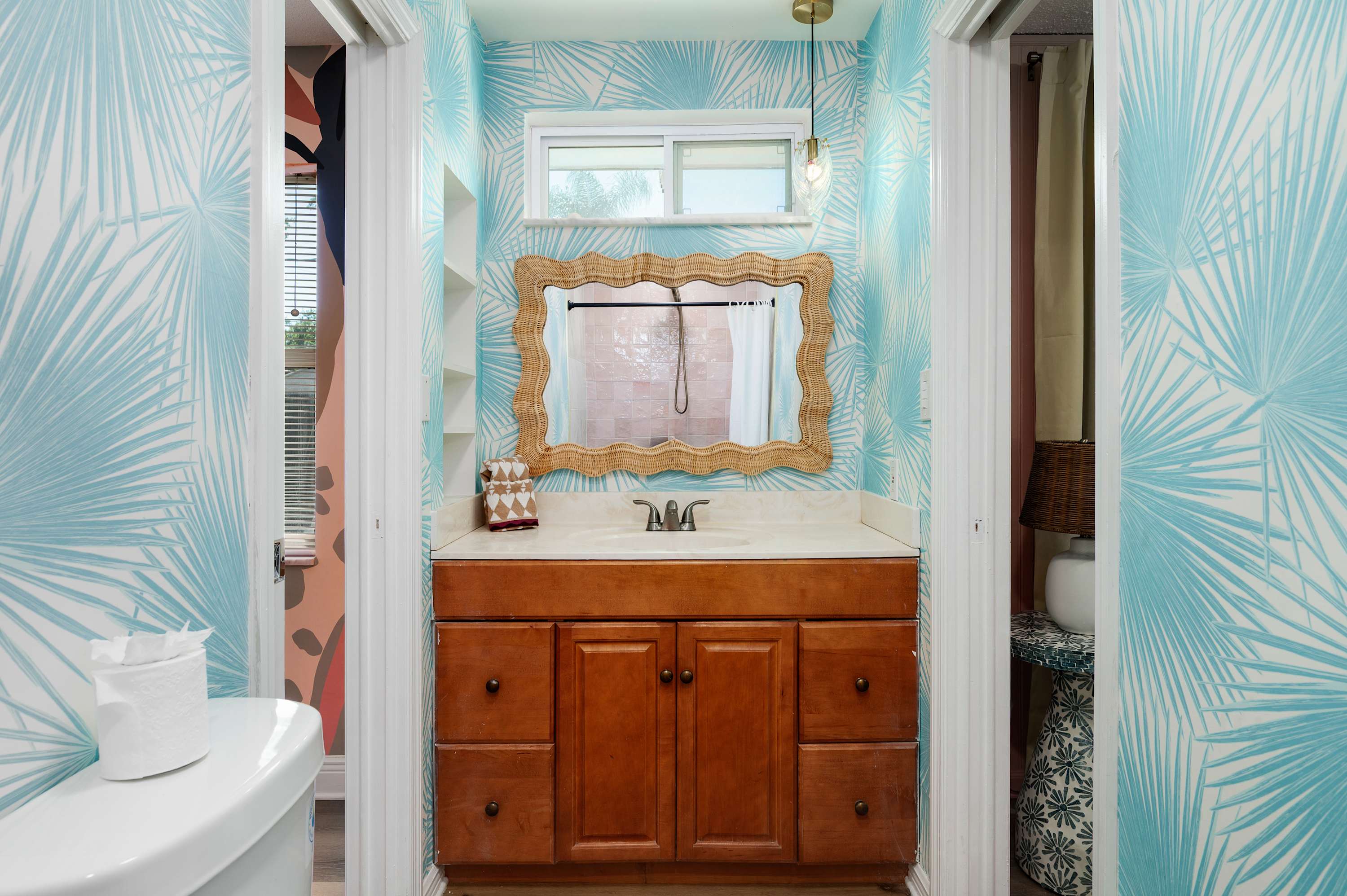 Bathroom with tropical leaf wallpaper and a wicker-framed mirror above the wooden vanity.
