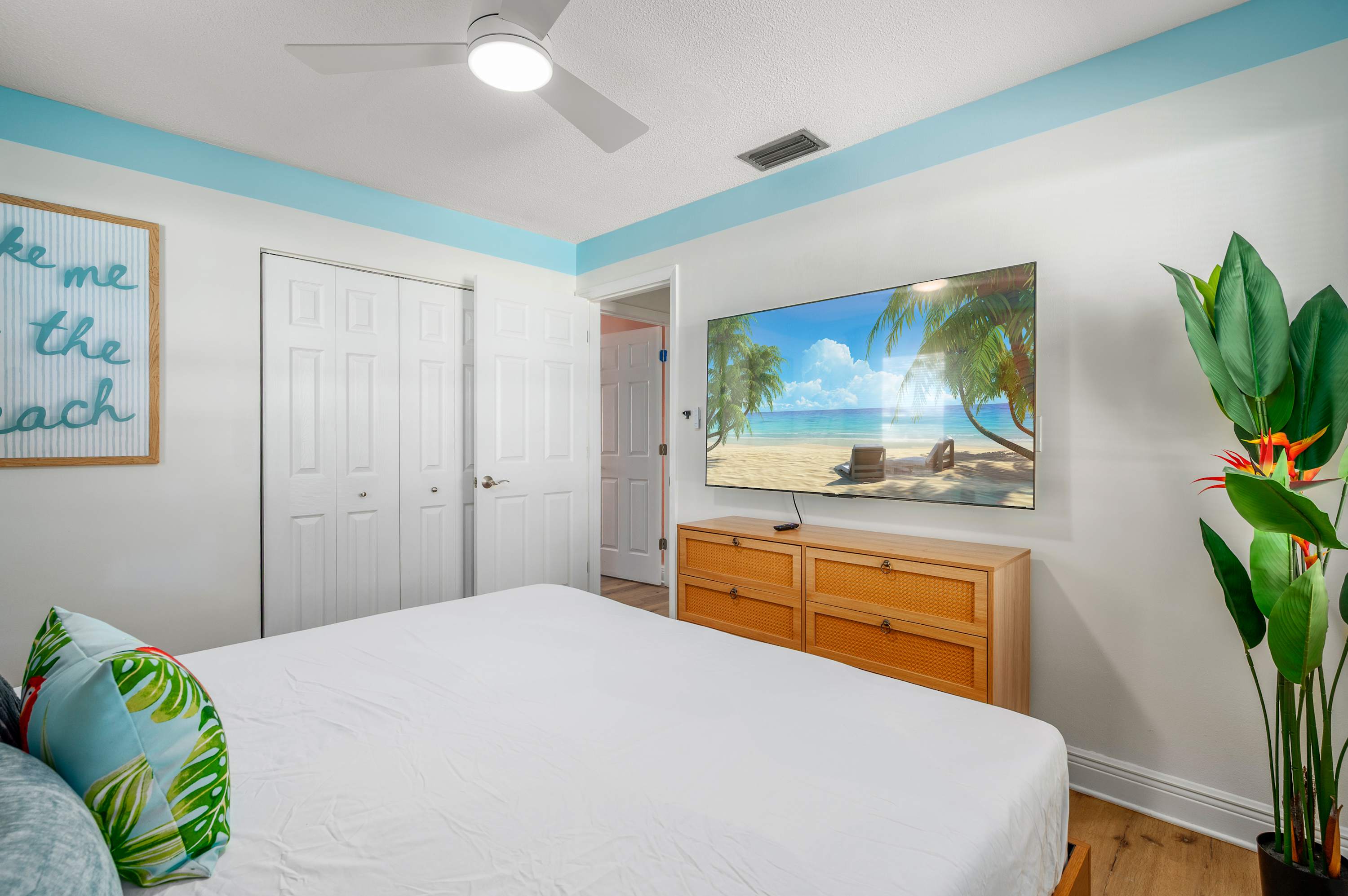 Relax in this bright bedroom featuring a ceiling fan and a large wall-mounted TV with tropical decor accents.