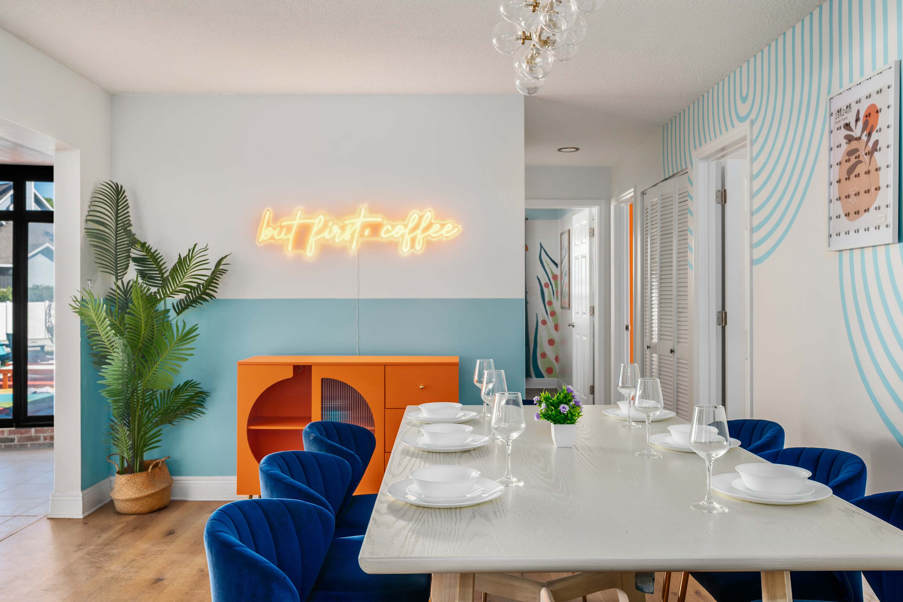 Dining area with a modern white table, blue velvet chairs, and a neon sign that reads 'but first coffee'.
