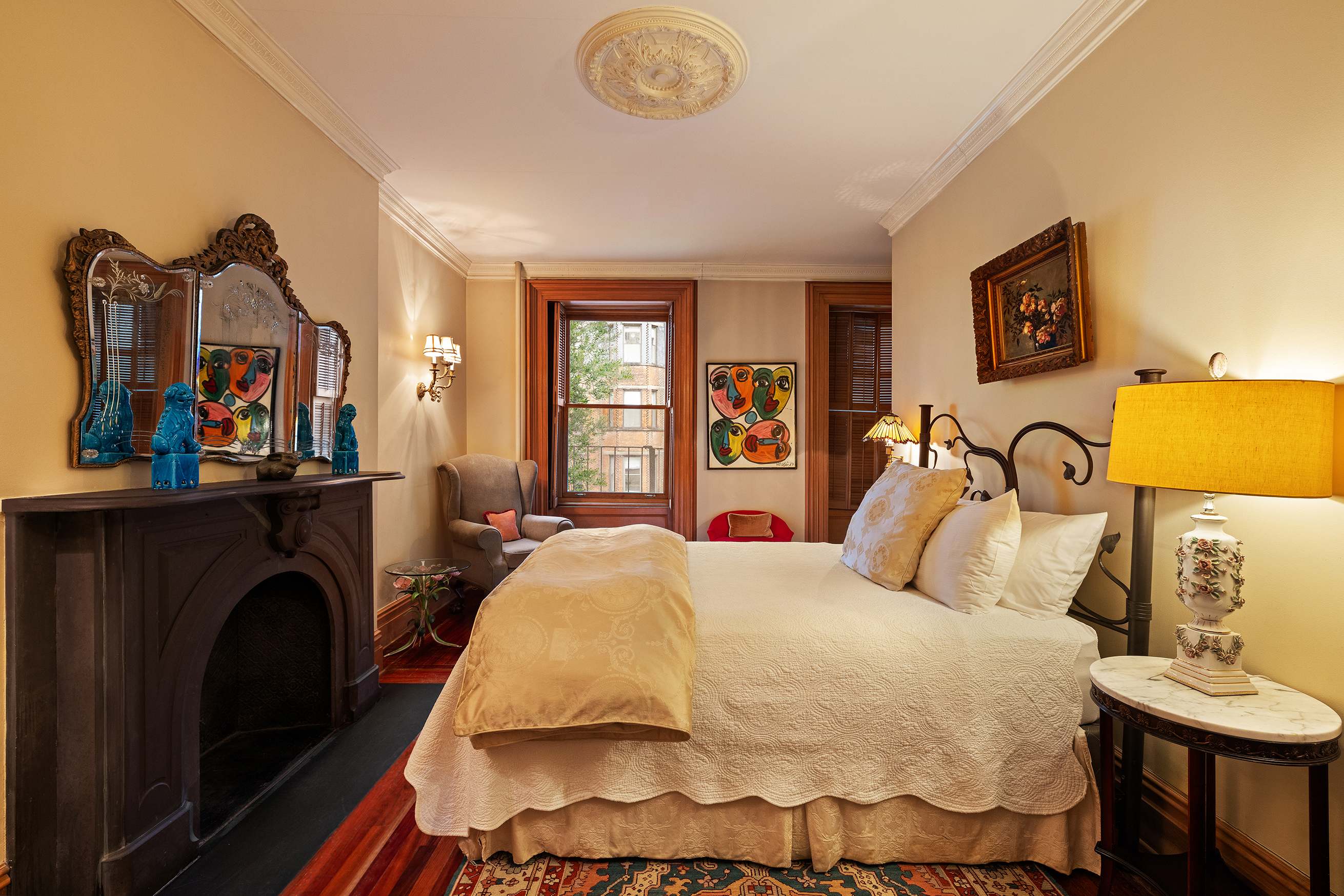 A wide view of a guest room with a bed, fireplace, window, and artwork.