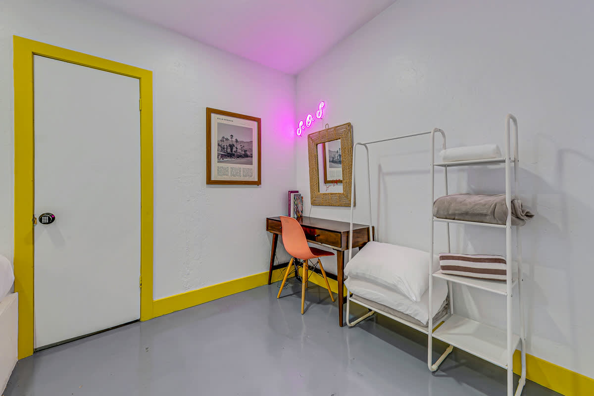 Dressing area with a small desk, chair, open clothing rack and yellow-trimmed door.