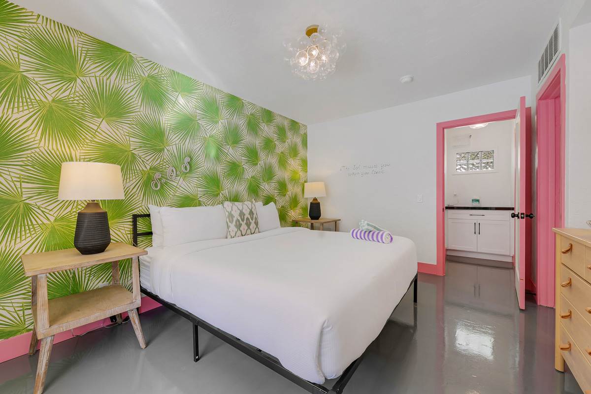 White bed against a green patterned accent wall with pink bedside furniture.