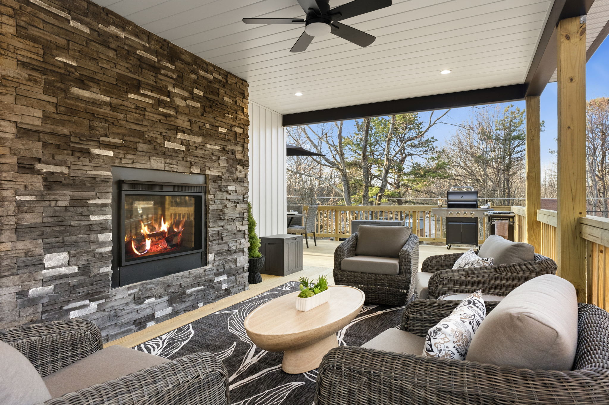 Covered porch lounge with stone fireplace, ceiling fan, and forest views