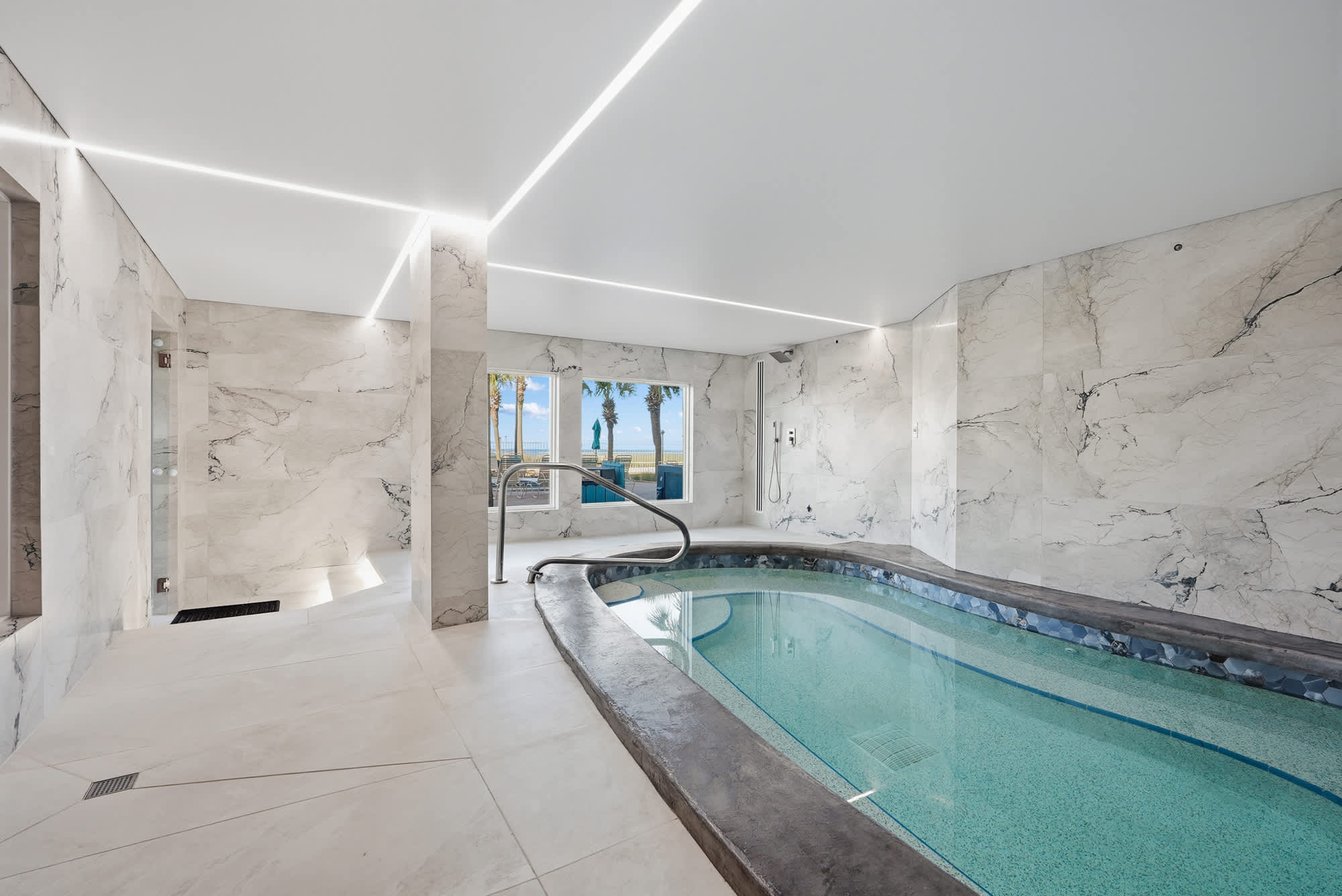 The Summit 1208 – Indoor pool area with marble walls and large windows overlooking palm trees and a beach view.