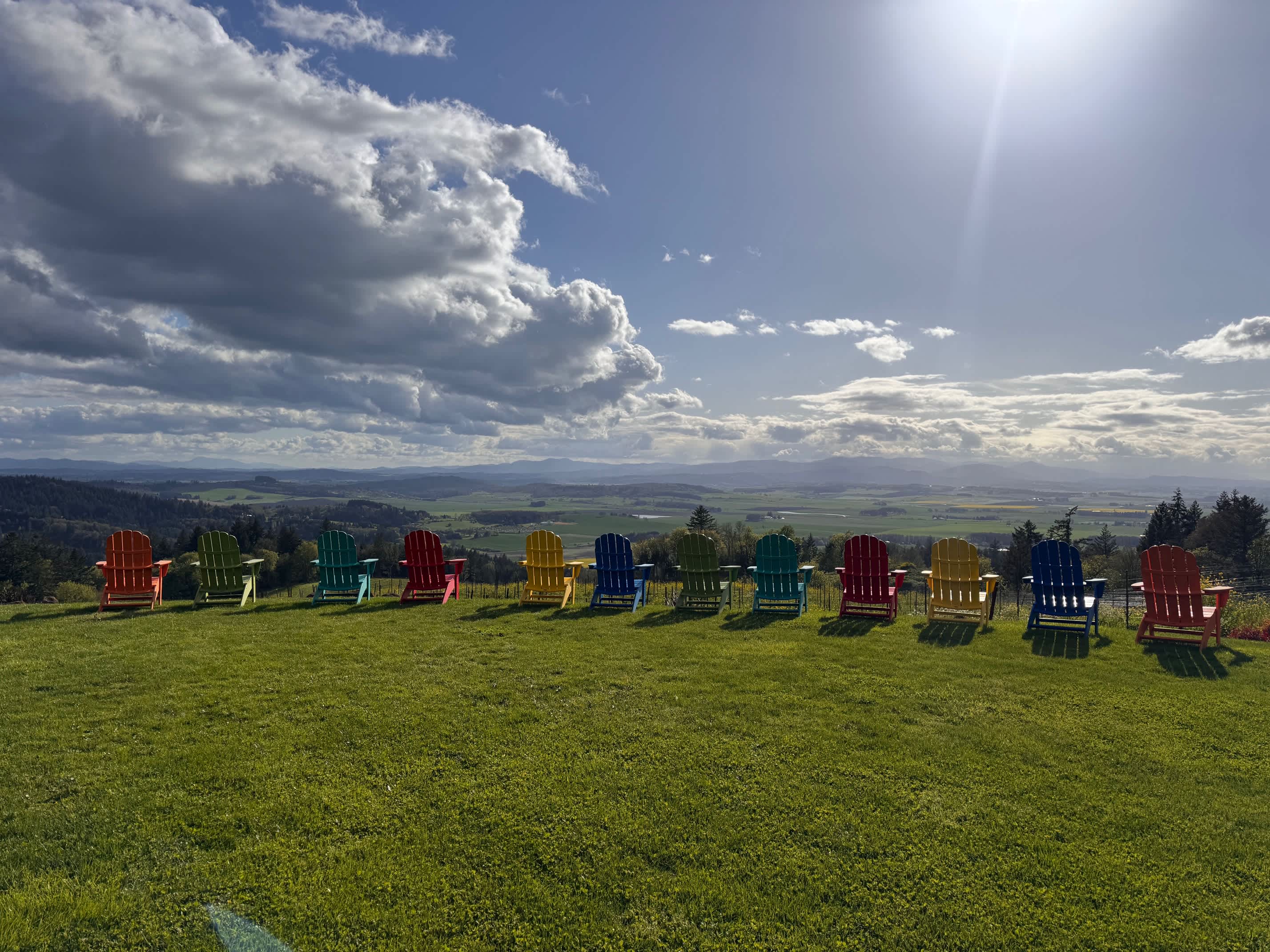 A line of vibrant, multi-colored Adirondack chairs arranged on a grassy hilltop, offering panoramic views of a sprawling valley and distant mountains under a bright, partly cloudy sky. The sun shines brightly from the upper right.