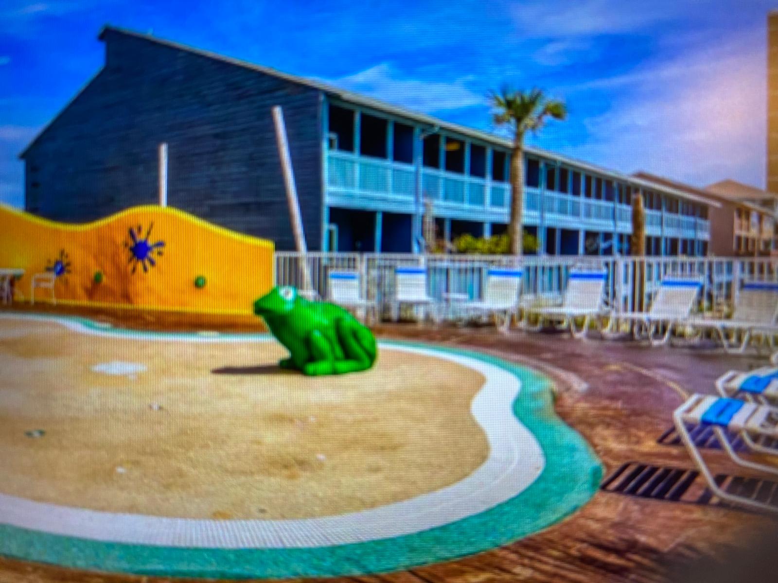 Splash 804E – Swimming pool area with a green frog statue and lounge chairs, behind a blue-painted building.