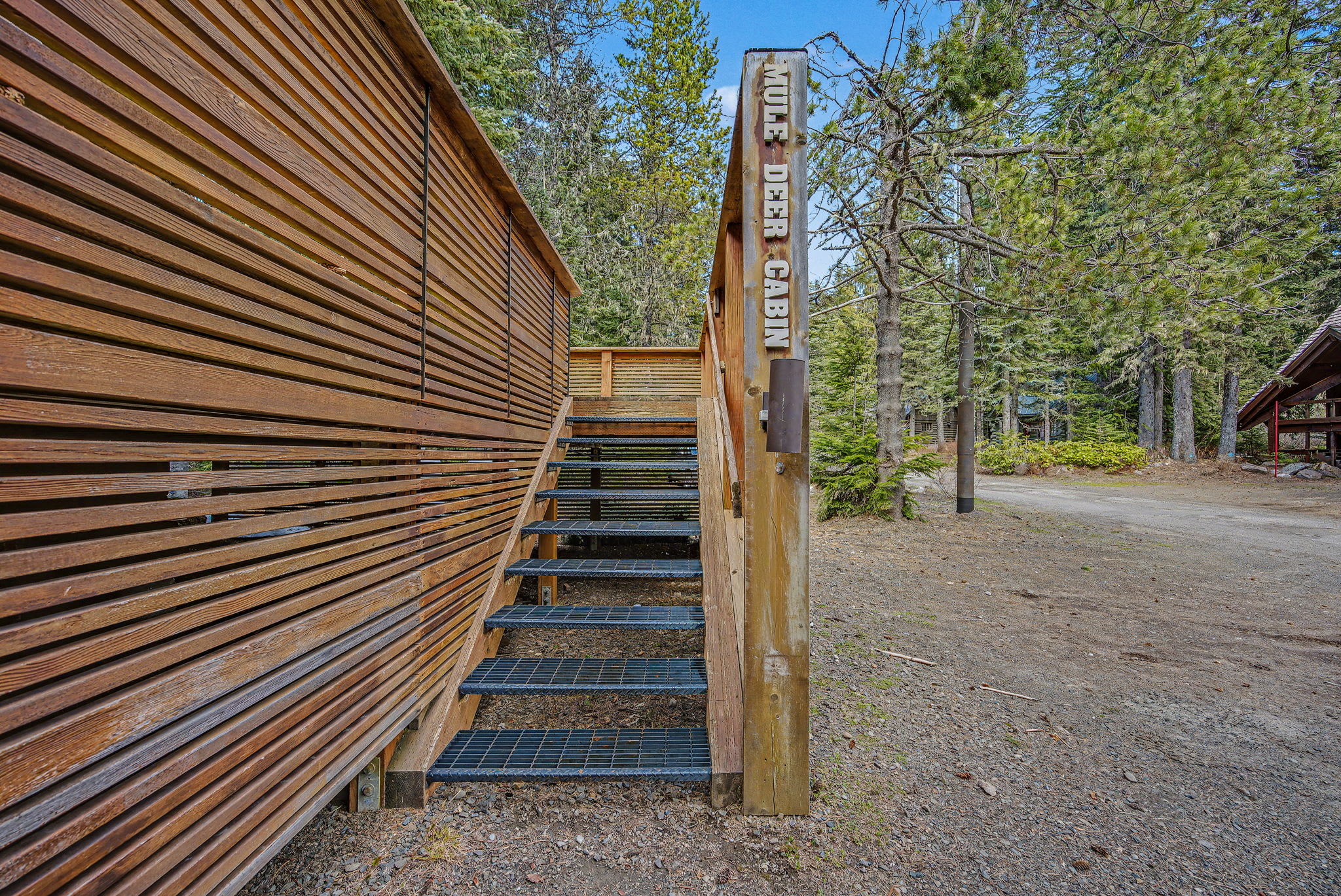An outdoor metal staircase with mesh treads leads up to the main entrance, flanked by a slatted wooden wall. A sign on the post reads 'MULE DEER CABIN'.