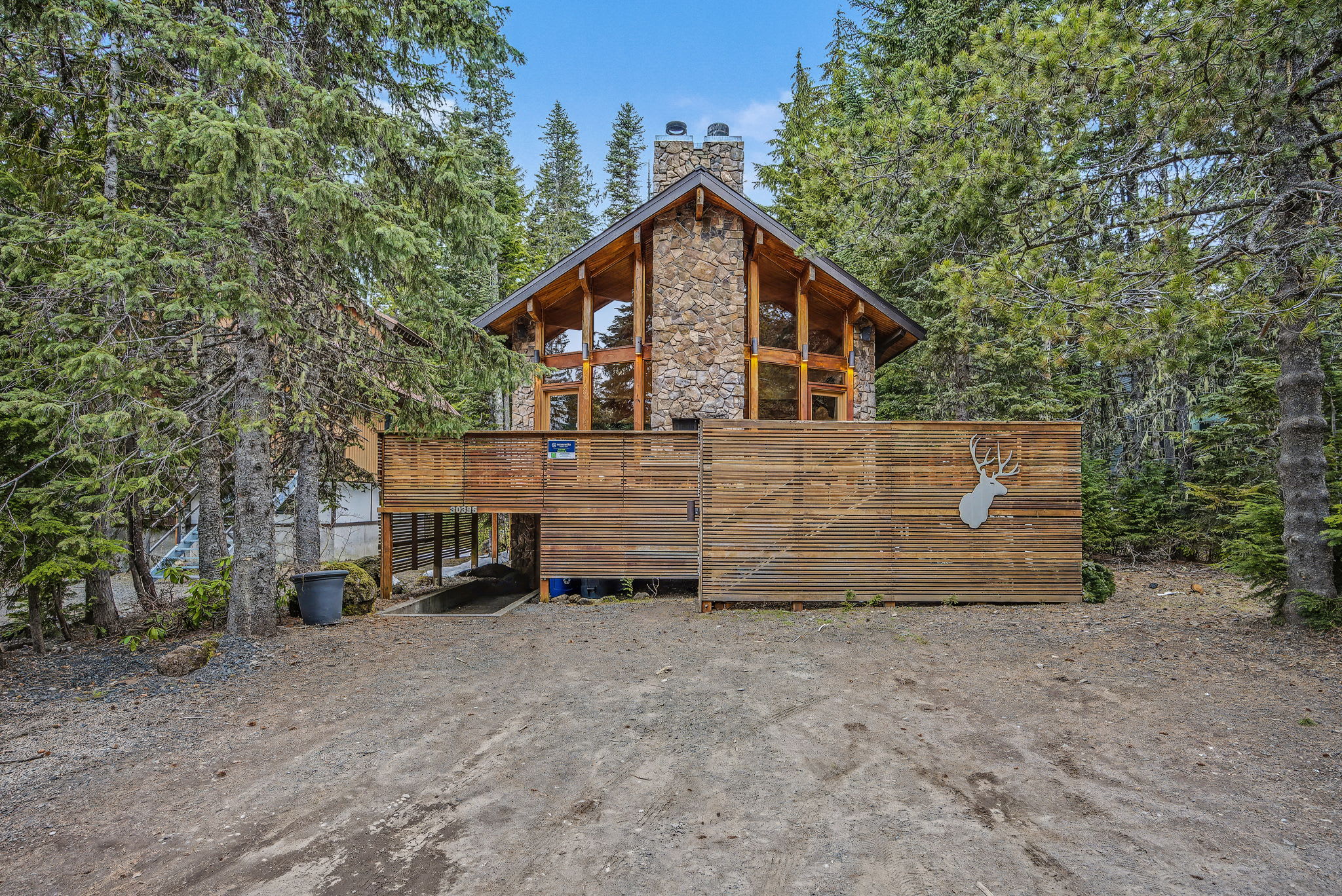 The front exterior of the chalet, showcasing its stone and wood construction amidst a dense forest. A slatted wooden fence with a large white deer head cutout provides privacy and a distinctive entrance.