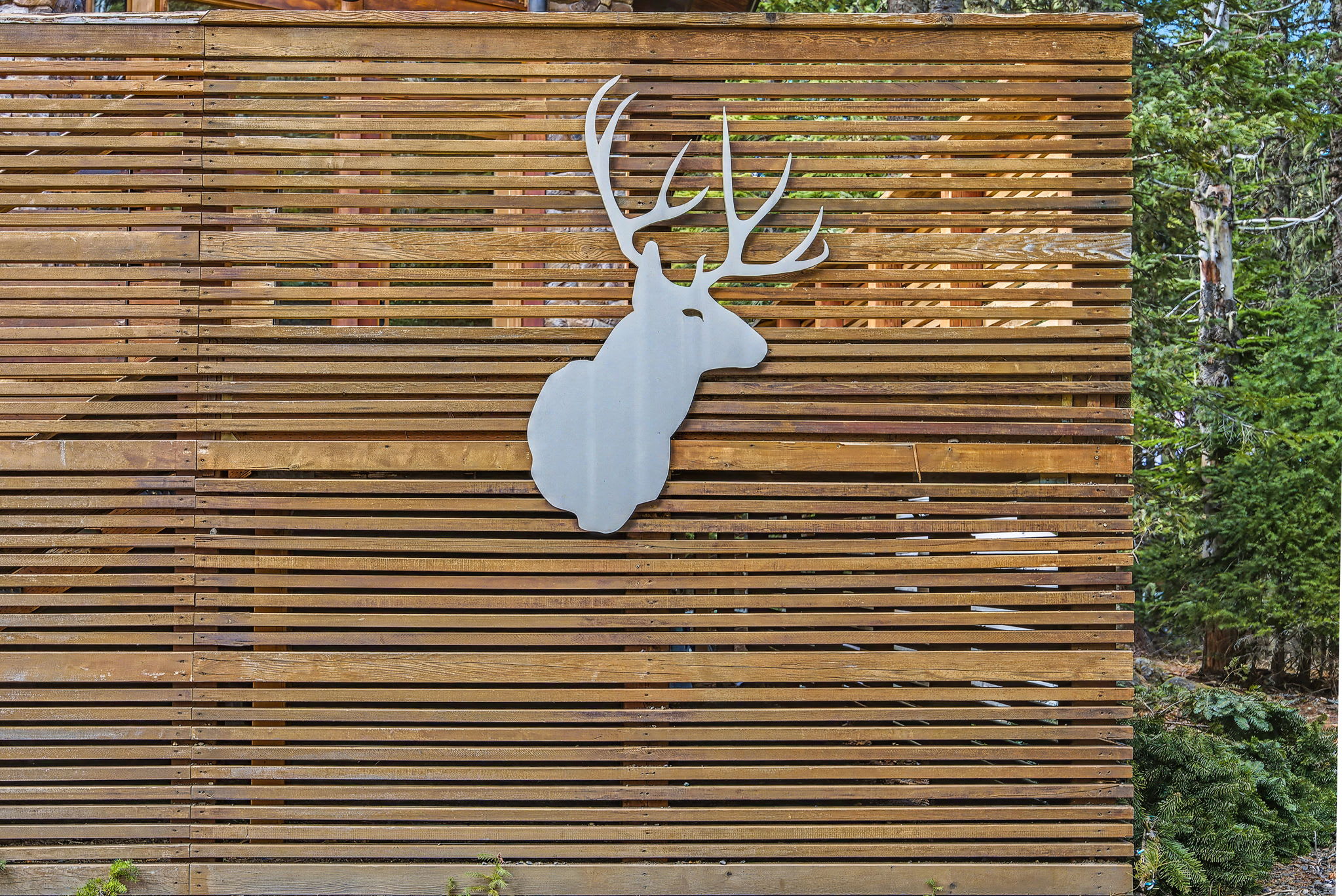 A close-up view of a large, white, stylized deer head cutout mounted on a horizontal slatted wooden fence, set against a backdrop of green trees.