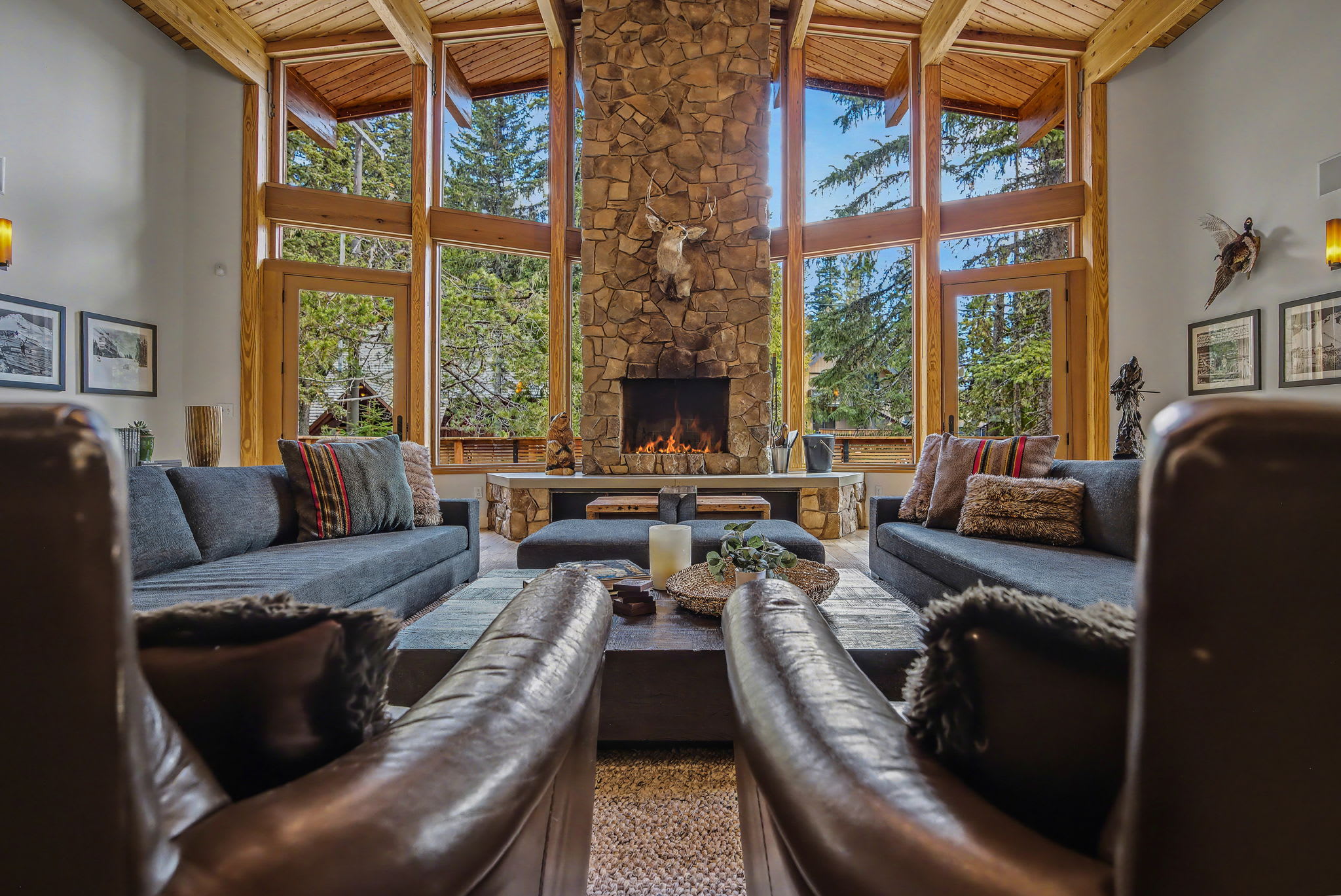 Grand living room with a towering stone fireplace as the focal point, flanked by two dark sofas. Floor-to-ceiling windows offer panoramic views of the surrounding forest and mountains.