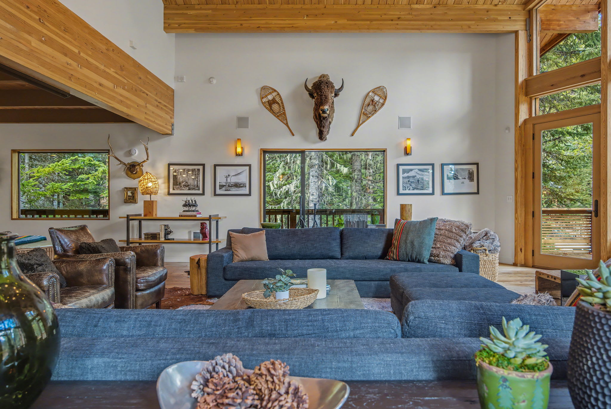 This wide view of the living room highlights a large dark sectional sofa, a wooden coffee table, and a wall decorated with a buffalo head mount and snowshoes. Large windows provide a lush green outdoor vista, enhancing the rustic-modern style.