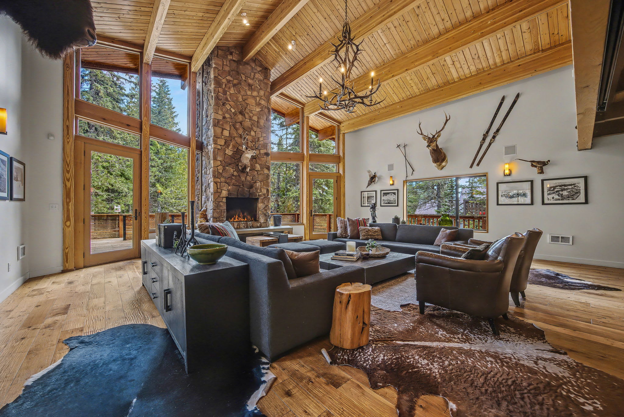 A grand living room with a soaring wood-paneled vaulted ceiling and exposed beams, centered around a massive stone fireplace. Multiple seating areas with dark sofas and leather armchairs are arranged on cowhide rugs, facing large windows with forest views.