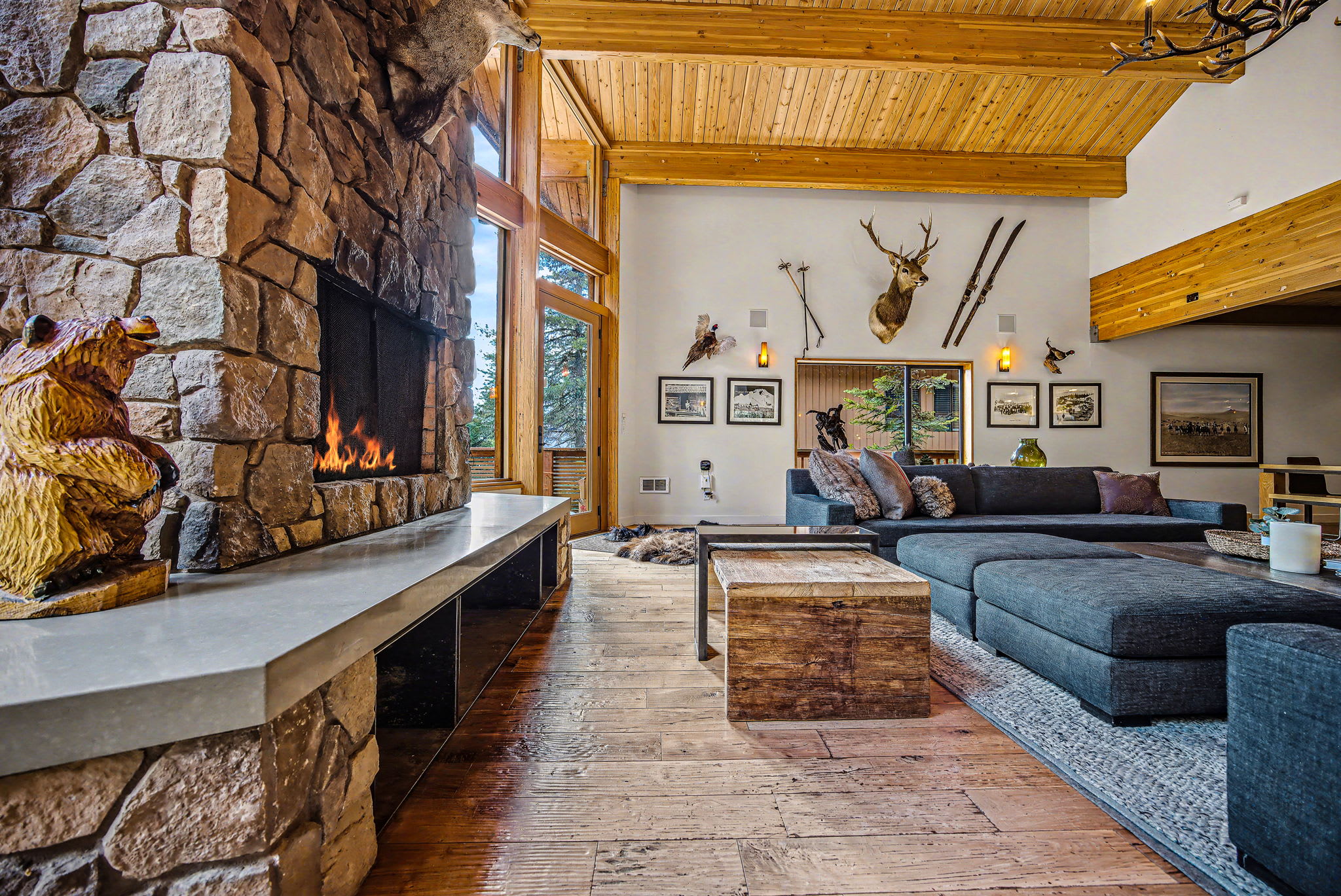 A cozy living room features a grand stone fireplace with a carved wooden bear statue, a dark sectional sofa, and a rustic wooden coffee table. Vaulted wooden ceilings and large windows offer views of the surrounding nature.