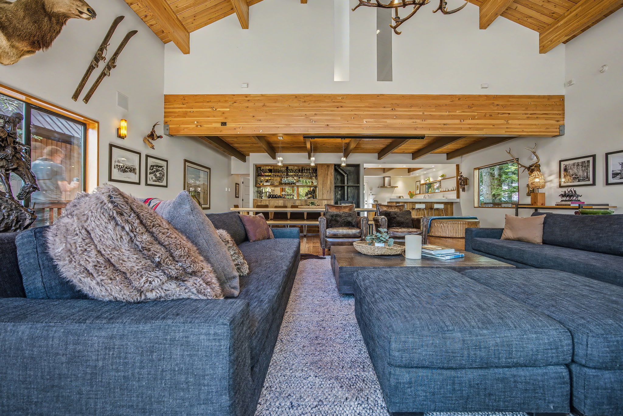 A spacious living room showcases a dark sectional sofa with plush pillows, hardwood floors, and a vaulted wooden ceiling. Animal mounts adorn the walls, and a built-in bar area with mirrored shelves is visible in the background.