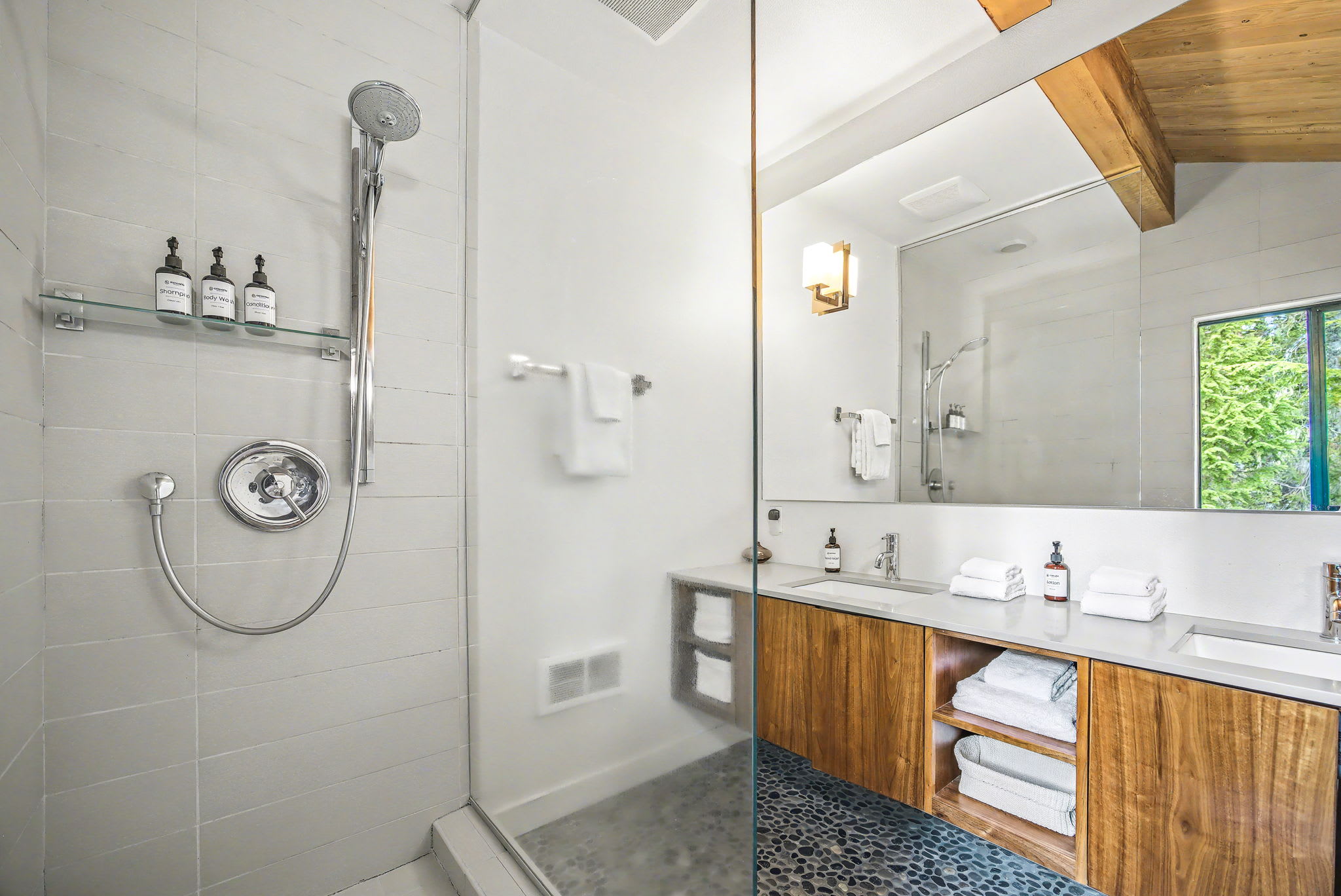 A close-up of the modern bathroom highlights the glass-enclosed shower with white tiled walls and a chrome rainfall showerhead. The wood vanity with a white countertop and dual sinks is reflected in the large mirror, complemented by dark pebble flooring and exposed wooden beams.