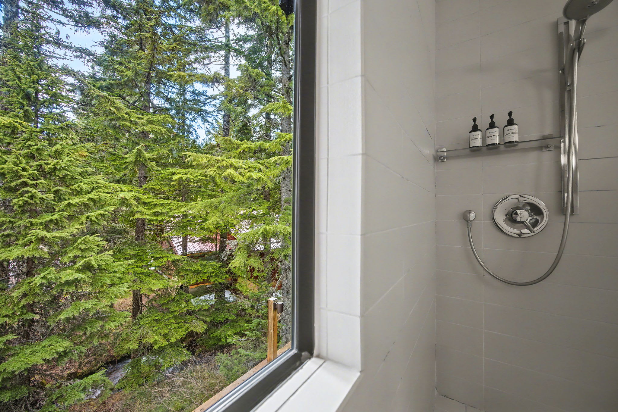 A large, modern window within the shower offers a stunning, immersive view of vibrant green pine trees and lush forest outside. The shower features white tiled walls and a chrome showerhead with wall-mounted dispensers.