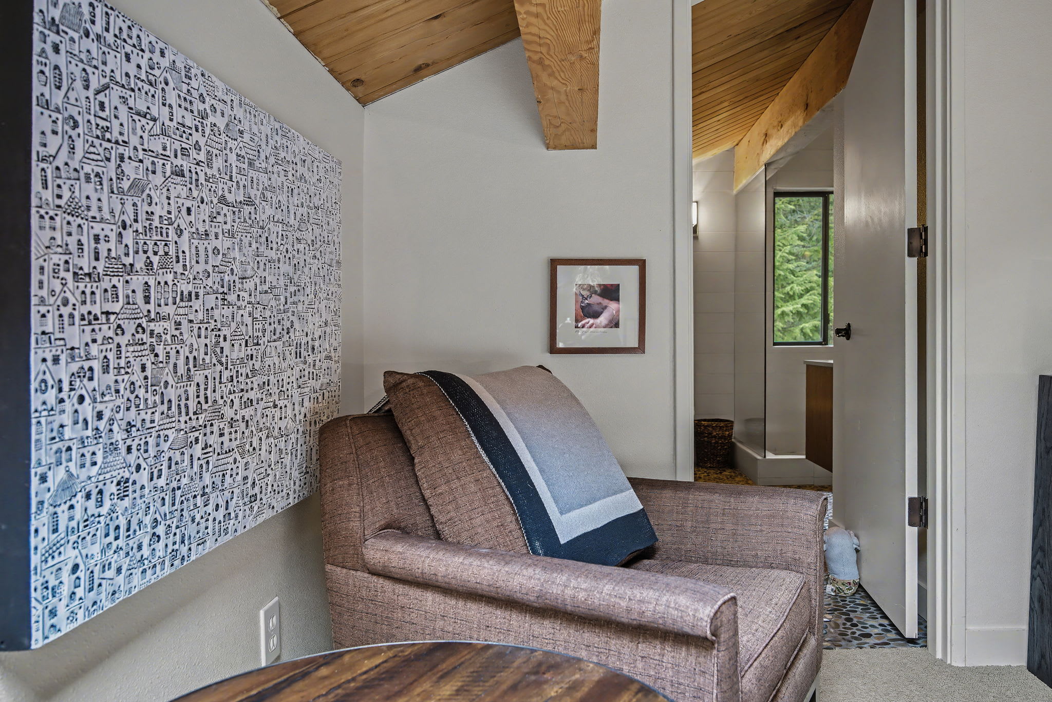 A cozy reading nook features a brown upholstered armchair with a striped blanket, positioned against a white wall adorned with a large black-and-white patterned art piece. A framed picture hangs above the chair, and a wooden table is partially visible, with an open doorway leading to a bathroom on the right.