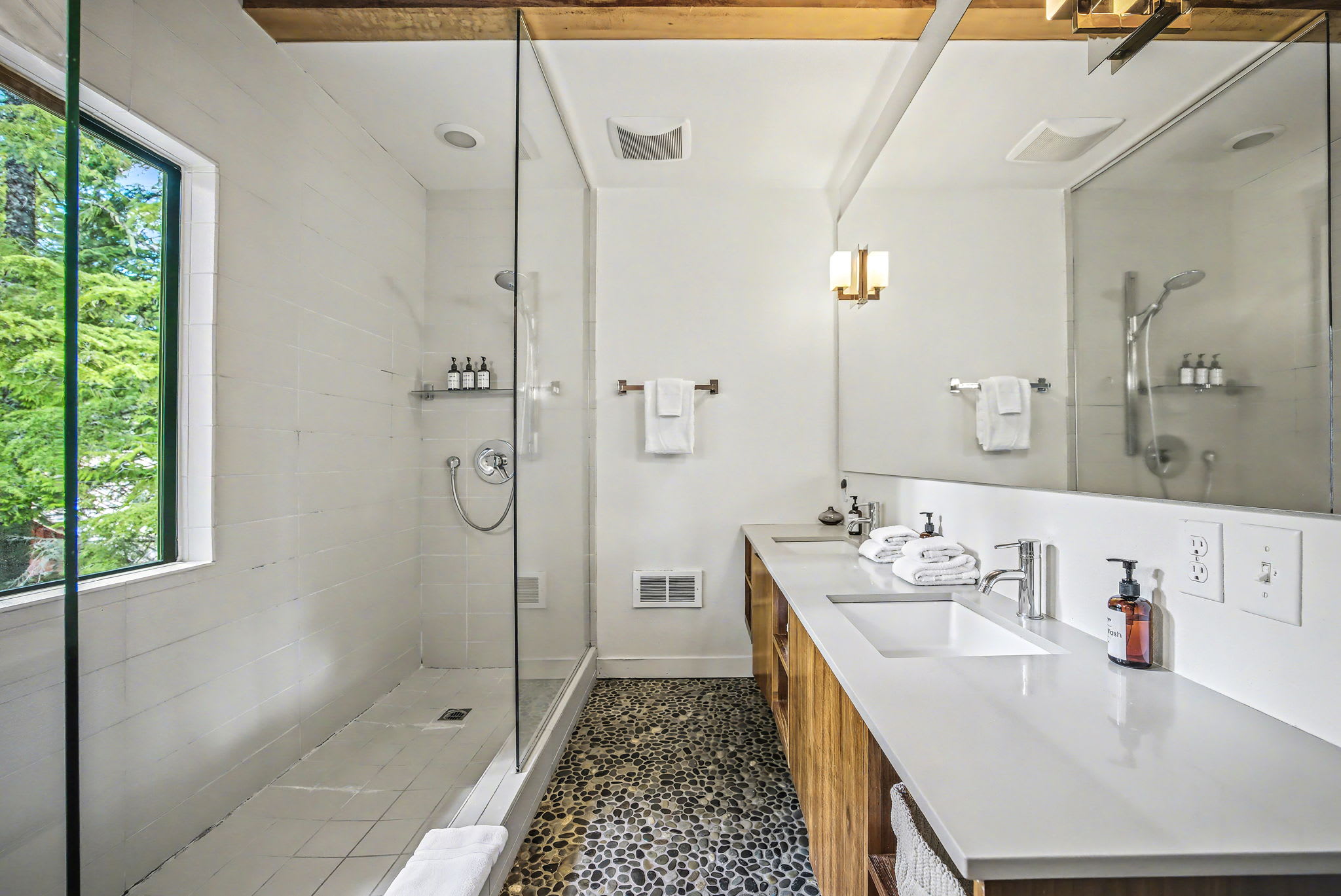 A bright, modern bathroom features a spacious glass-enclosed shower with white subway tiles and a large window offering lush green tree views. A double vanity with a white countertop, wood cabinetry, and dark pebble flooring completes the contemporary design under exposed wooden beams.