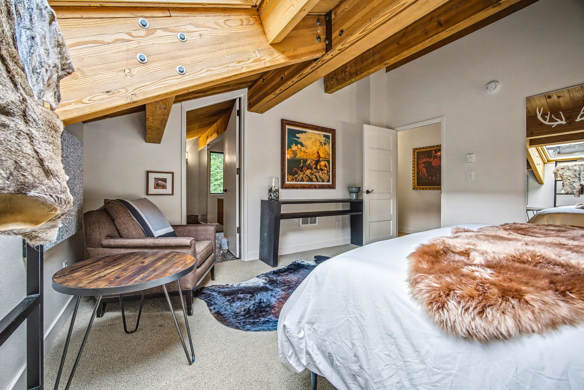 Another view of the attic bedroom, showing a cozy armchair and a small side table. The sloped ceiling with exposed wooden beams and a skylight are prominent features, adding to the rustic charm.