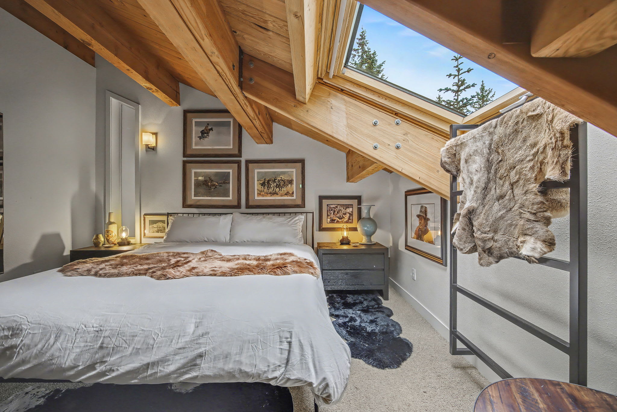 Attic bedroom with a queen-sized bed, white bedding, and a faux fur throw. The room features a sloped wood-paneled ceiling with exposed beams and a large skylight offering views of the sky.