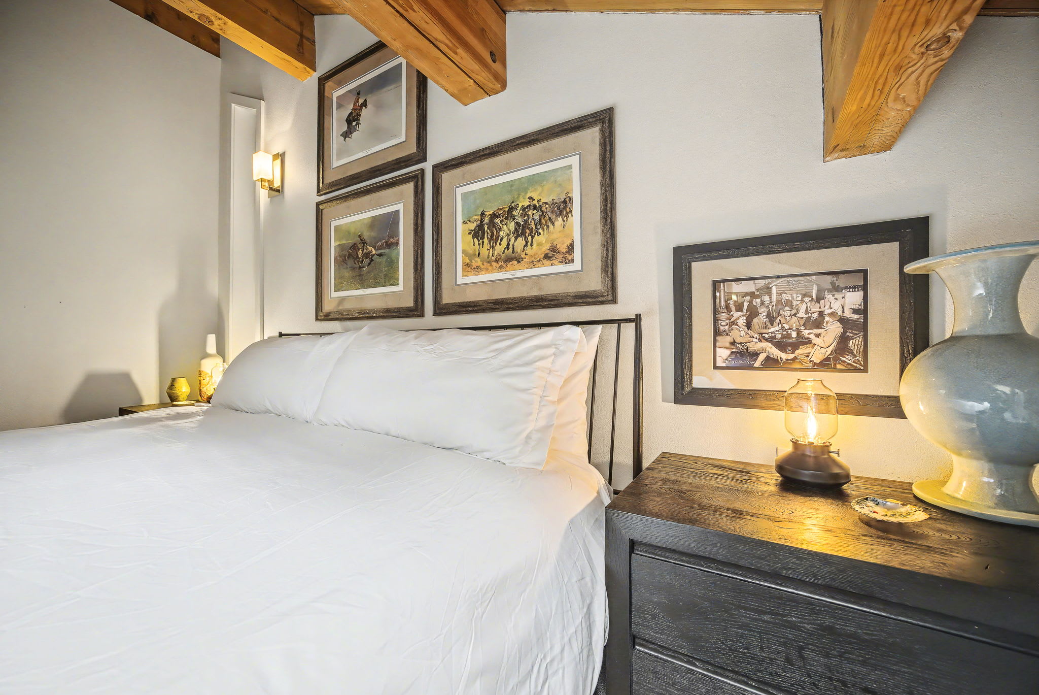 A close-up of the bedroom highlights the white-linened bed with a dark metal headboard, flanked by a dark nightstand with a decorative lantern. Multiple framed artworks hang on the white wall, beneath the rustic exposed wooden beams of the vaulted ceiling.