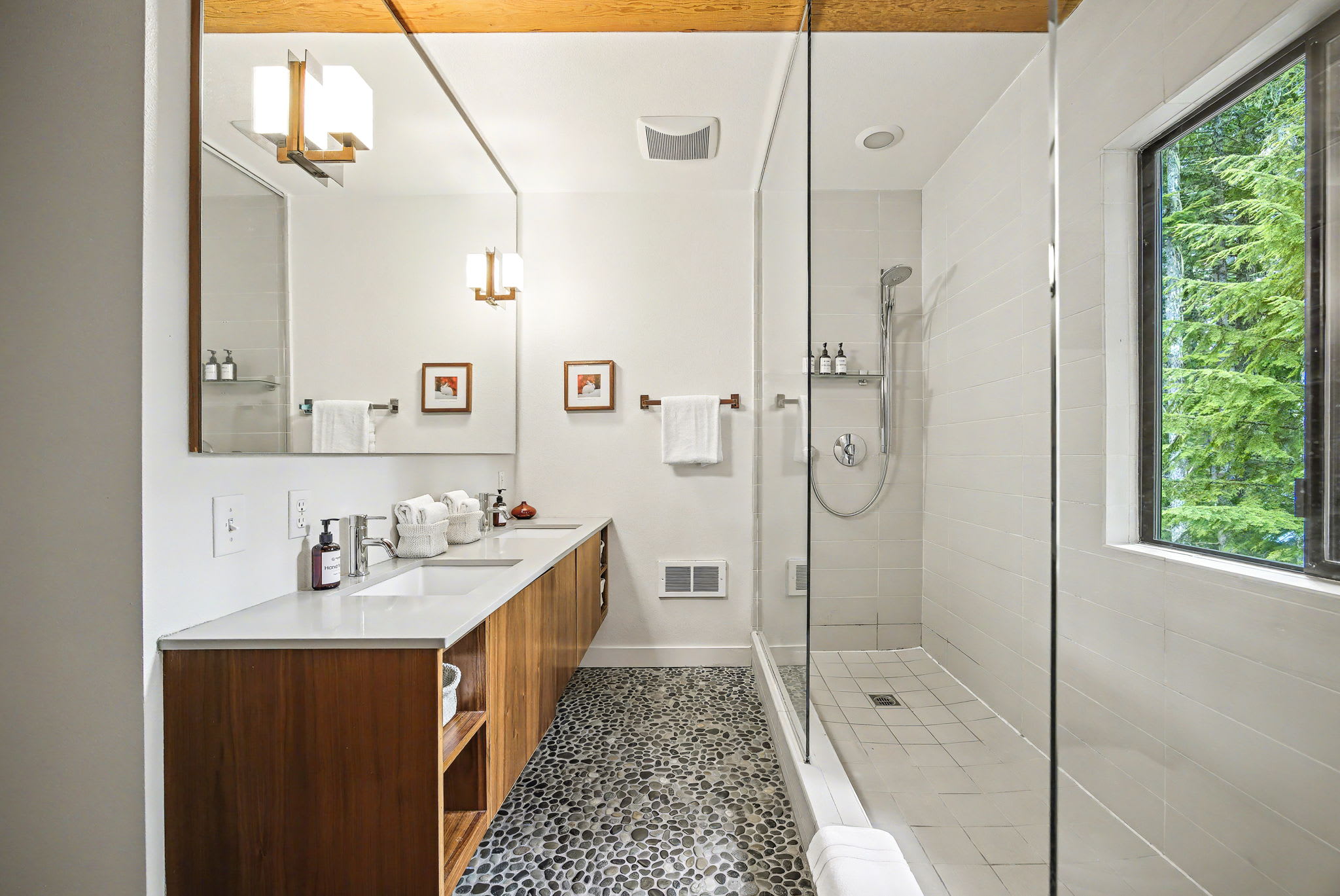 A modern bathroom featuring a double vanity with a wooden cabinet and white countertop, a large mirror, and a spacious glass-enclosed shower. The room is set against a unique pebble-stone floor and a warm wooden beam ceiling.