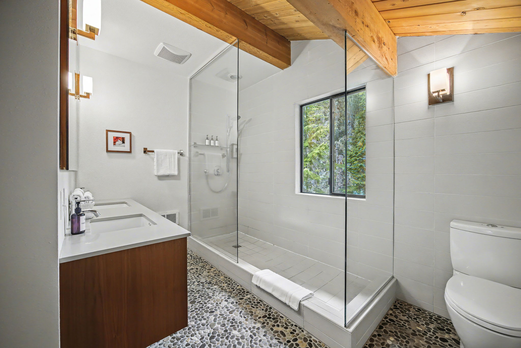 A bright bathroom with a glass-enclosed shower and a modern toilet, featuring a large window offering a serene view of lush green trees. The space is grounded by a pebble-stone floor and topped with a wooden beam ceiling.
