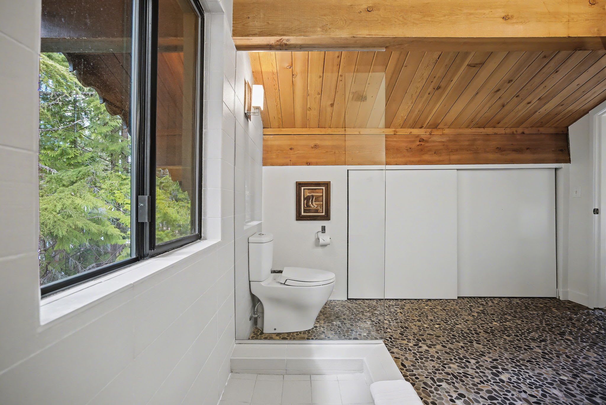 The toilet area of a bathroom, featuring a modern toilet and a large window overlooking a dense forest, creating a private and natural ambiance. The space is completed with a pebble-stone floor and a wooden beam ceiling.