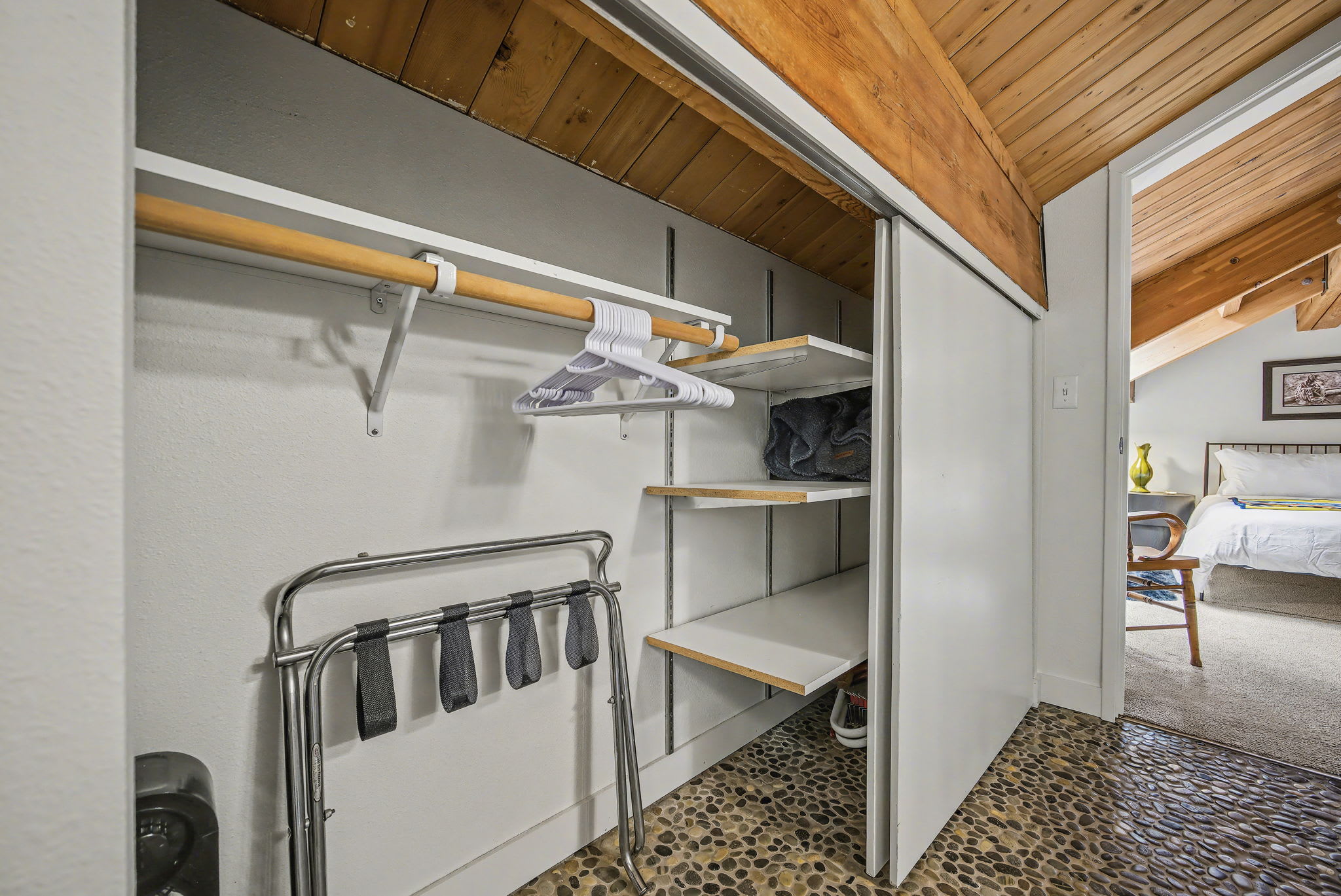 A well-organized closet area featuring a wooden hanging rod, white hangers, and open shelving, with a metal luggage rack on a unique pebble-stone floor. A glimpse of a bedroom with a bed is visible through the open doorway.